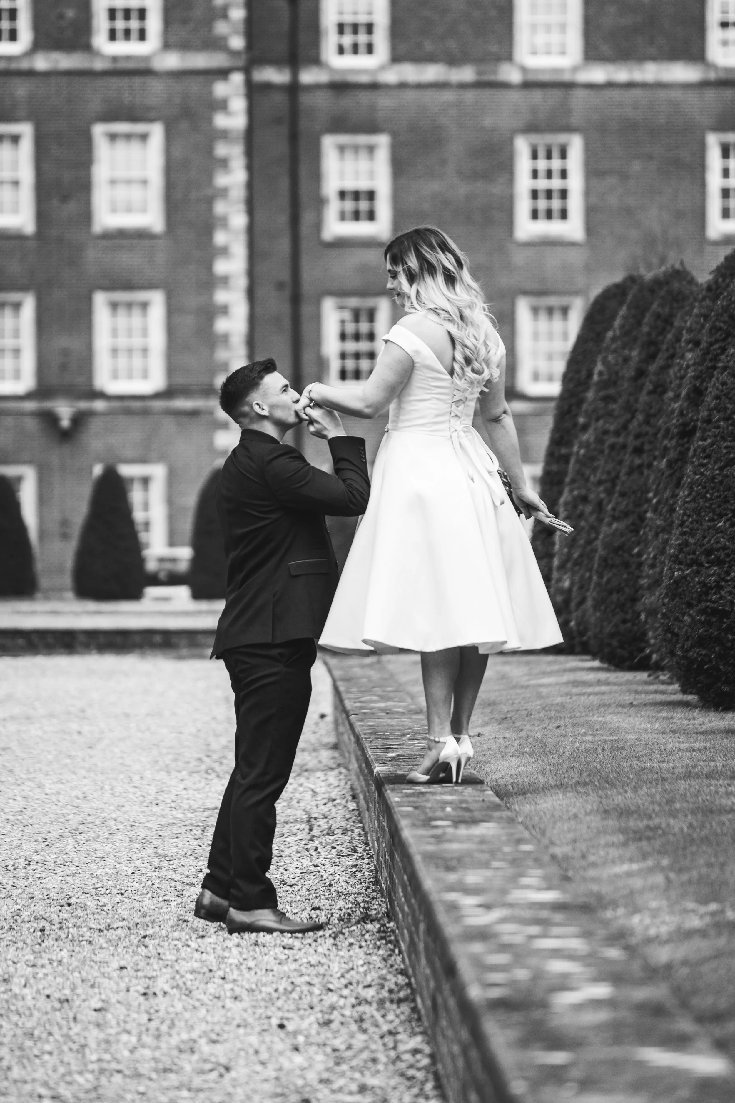A man kneeling and kissing a woman standing on a ledge, holding her hand, in front of a large brick building with windows and trimmed bushes, black and white photograph.