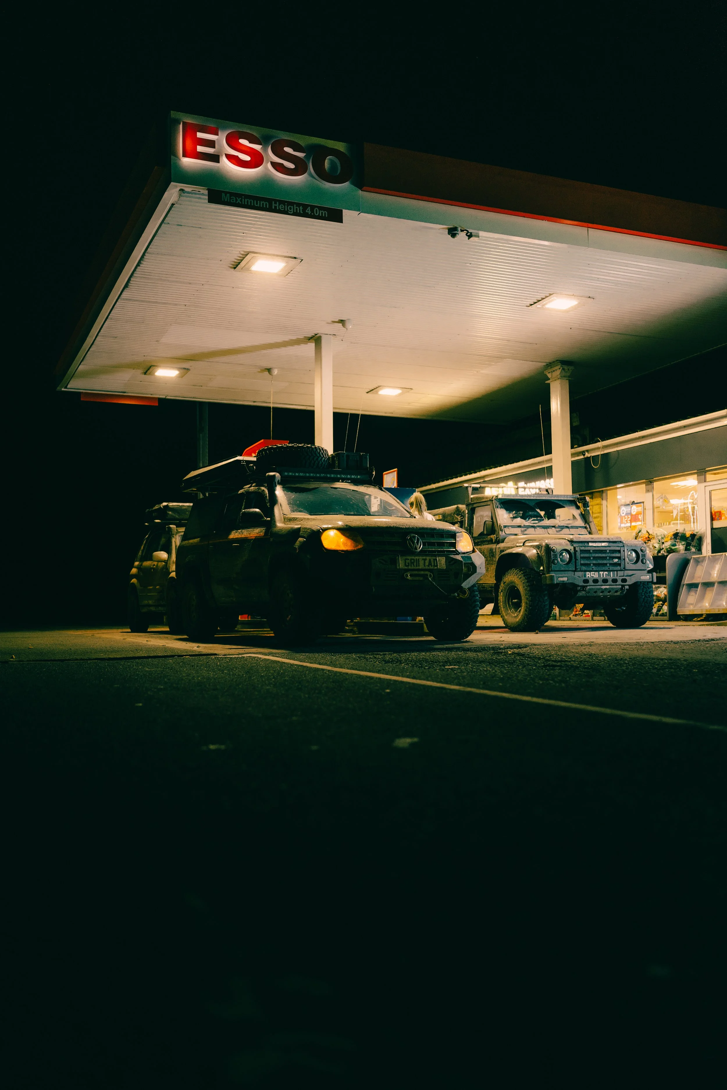 Night view of an ESSO gas station with several off-road vehicles parked, illuminated by overhead lights.