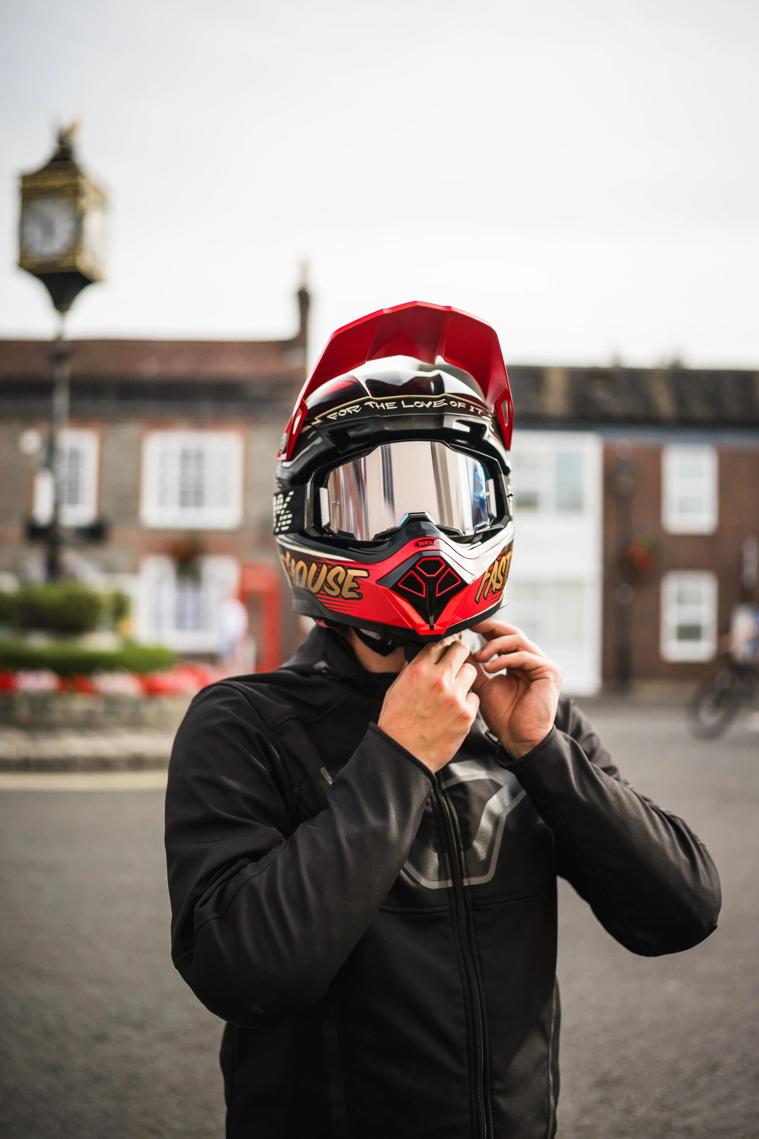 Person wearing a black jacket and a colorful full-face motorcycle helmet, standing outdoors in front of a building with a clock tower, adjusting the helmet strap.