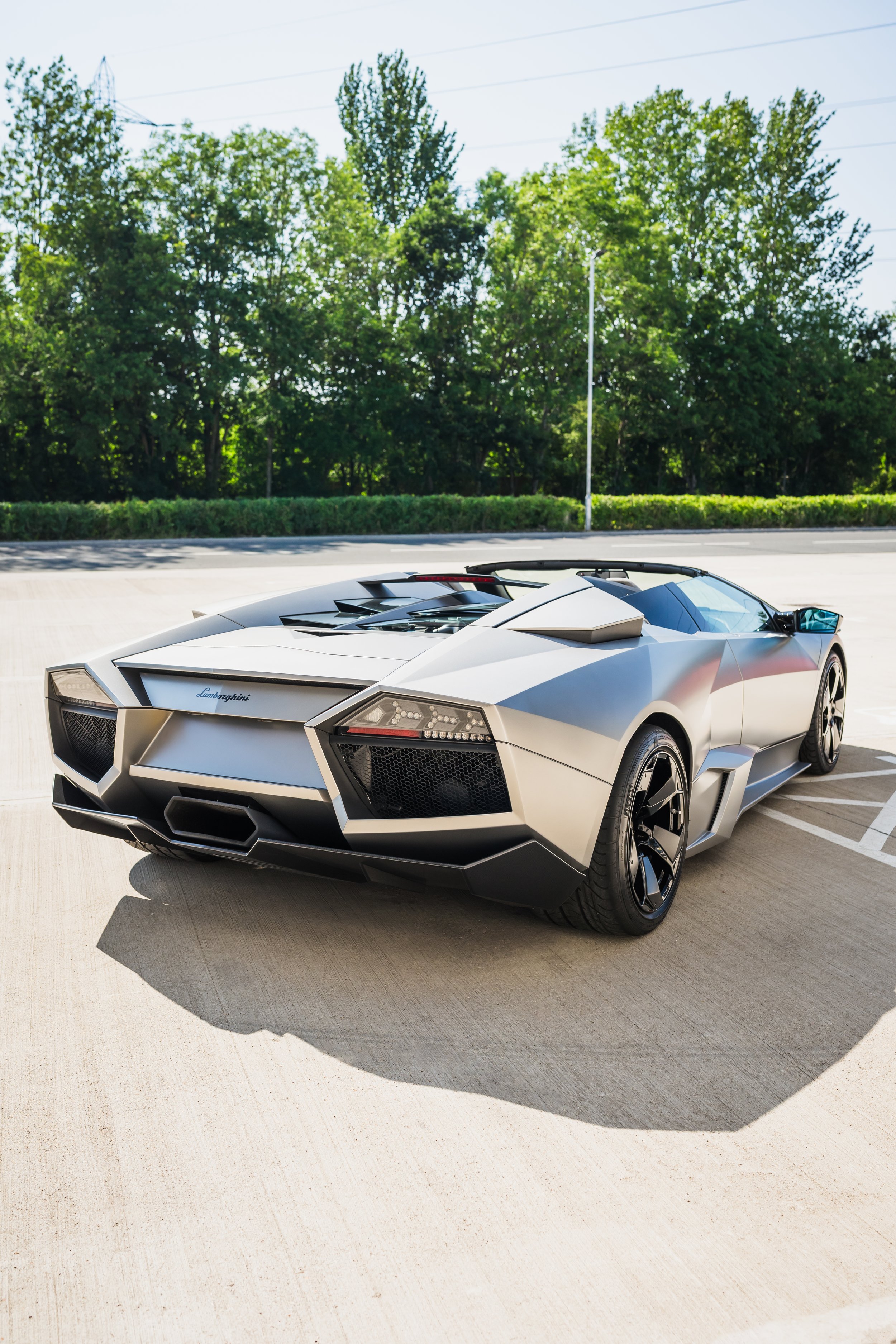 A silver Lamborghini sports car parked on a concrete parking lot with green trees and a blue sky in the background.