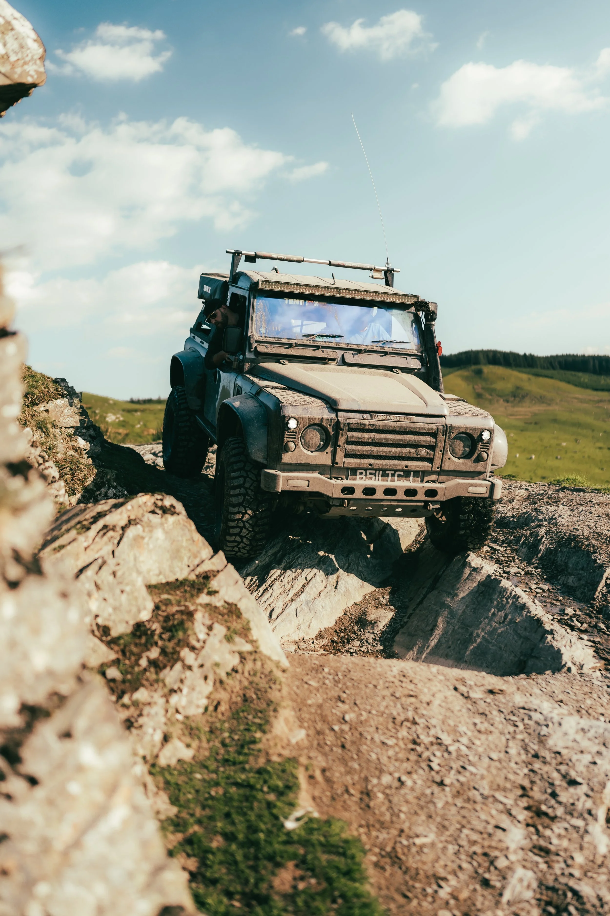A rugged off-road vehicle climbing over large rocks on a dirt trail in a mountainous landscape under a partly cloudy sky.
