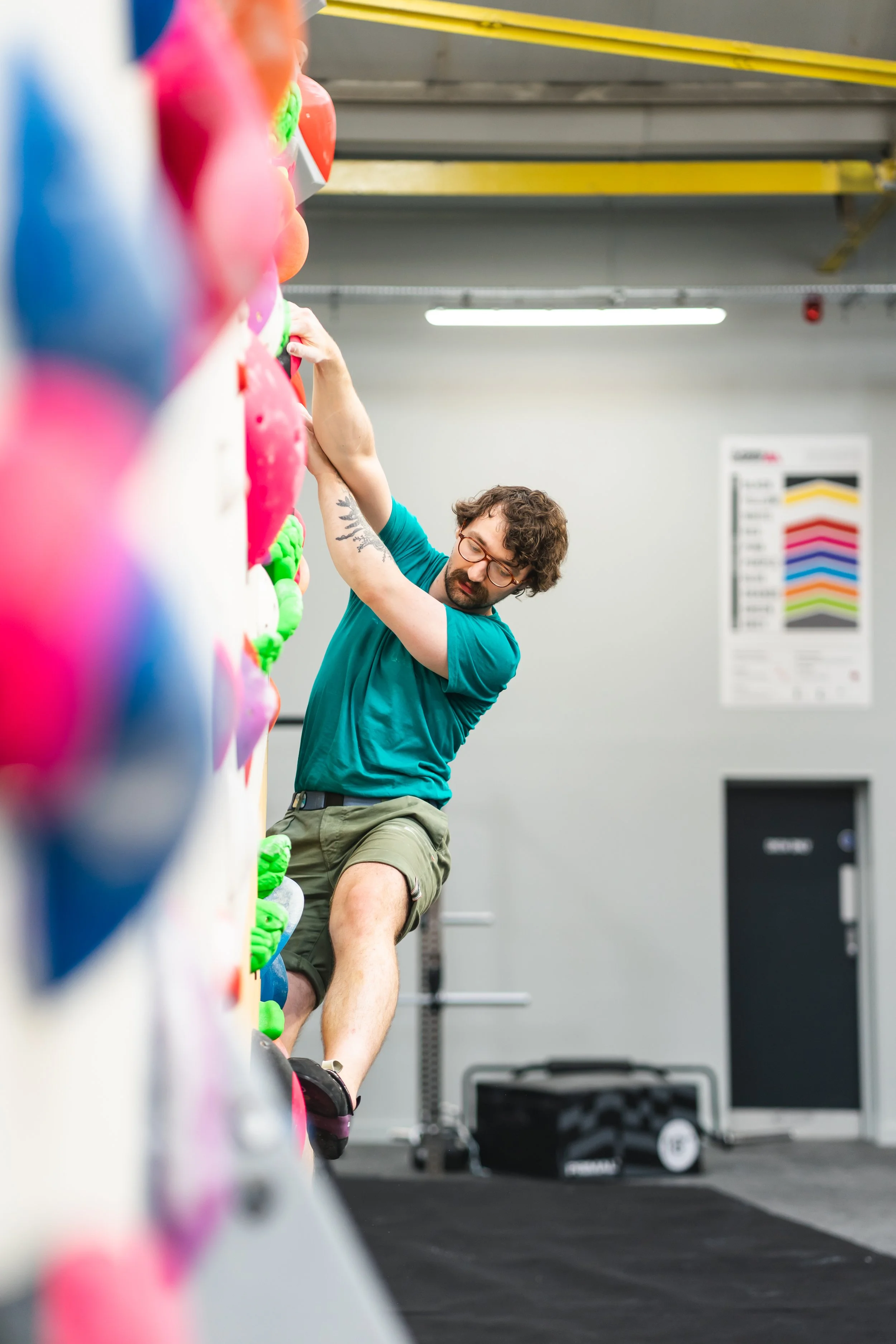 A man with glasses and curly hair climbing an indoor climbing wall with colorful holds.