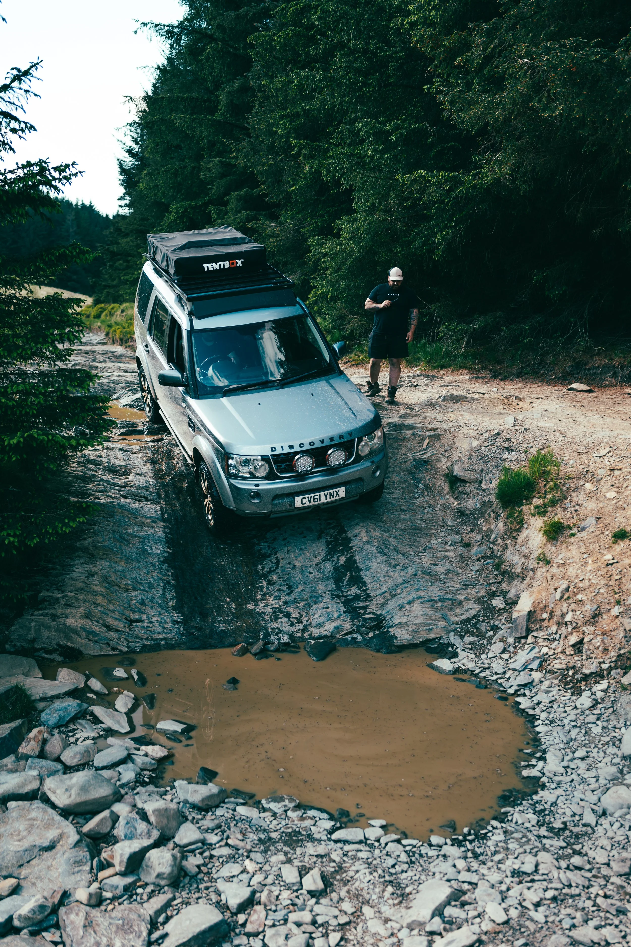 A silver Land Rover Discovery vehicle stuck in a muddy rut on a dirt trail, with a person standing nearby holding a phone and looking at it, surrounded by green trees.
