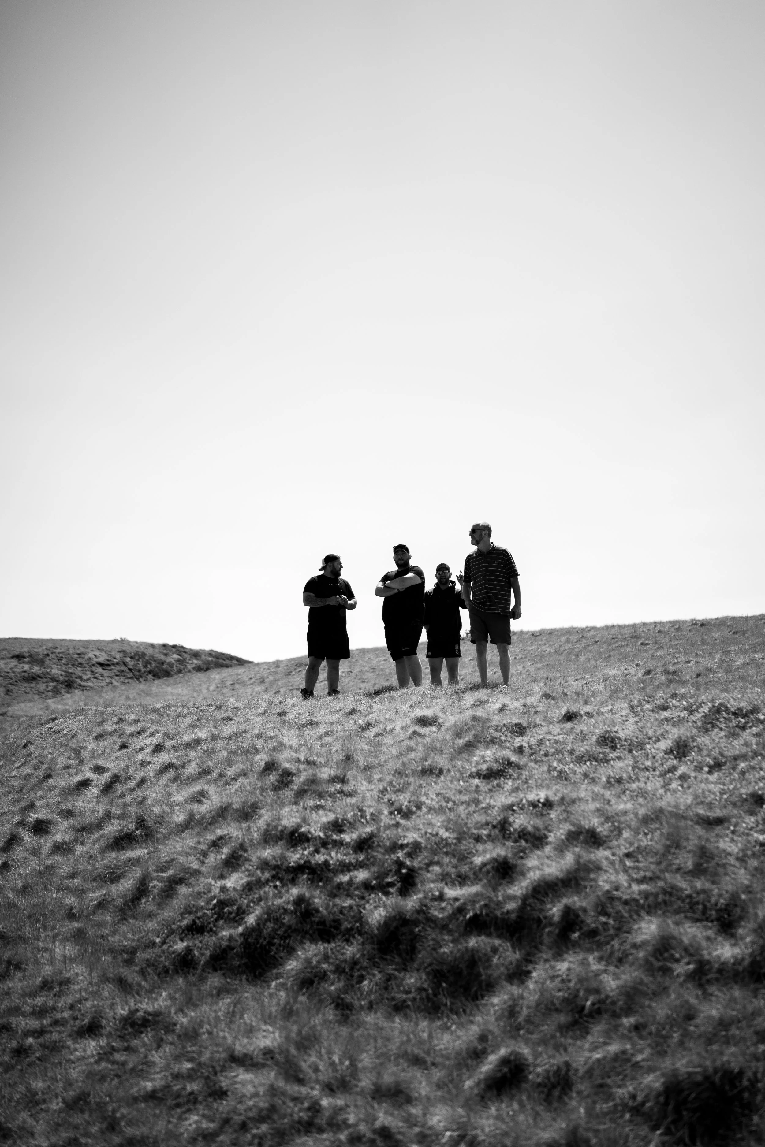 Four people standing on a grassy hill, silhouetted against the sky, with minimal clouds visible.