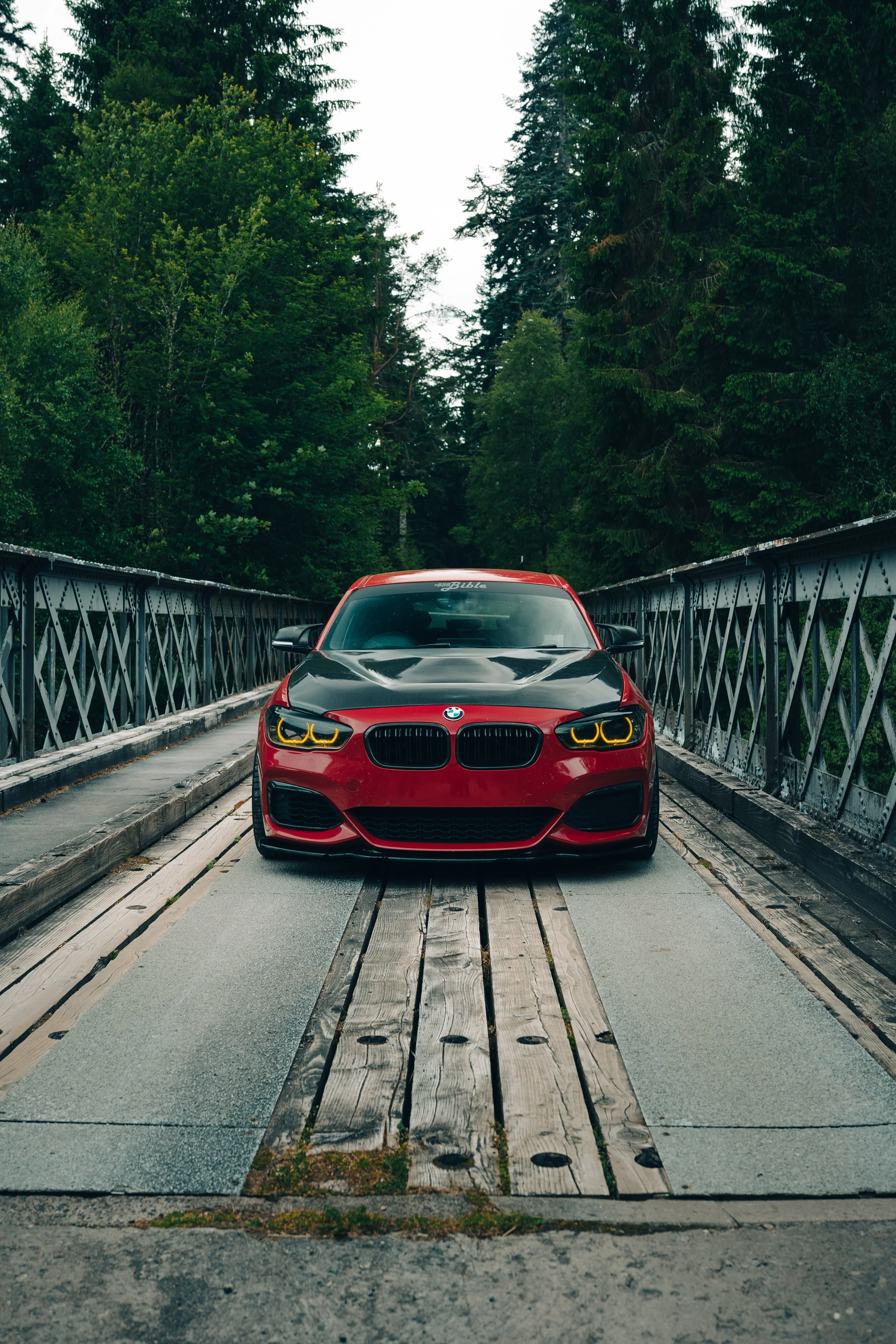 A red BMW car with black hood and yellow headlights parked on a wooden bridge surrounded by green trees.
