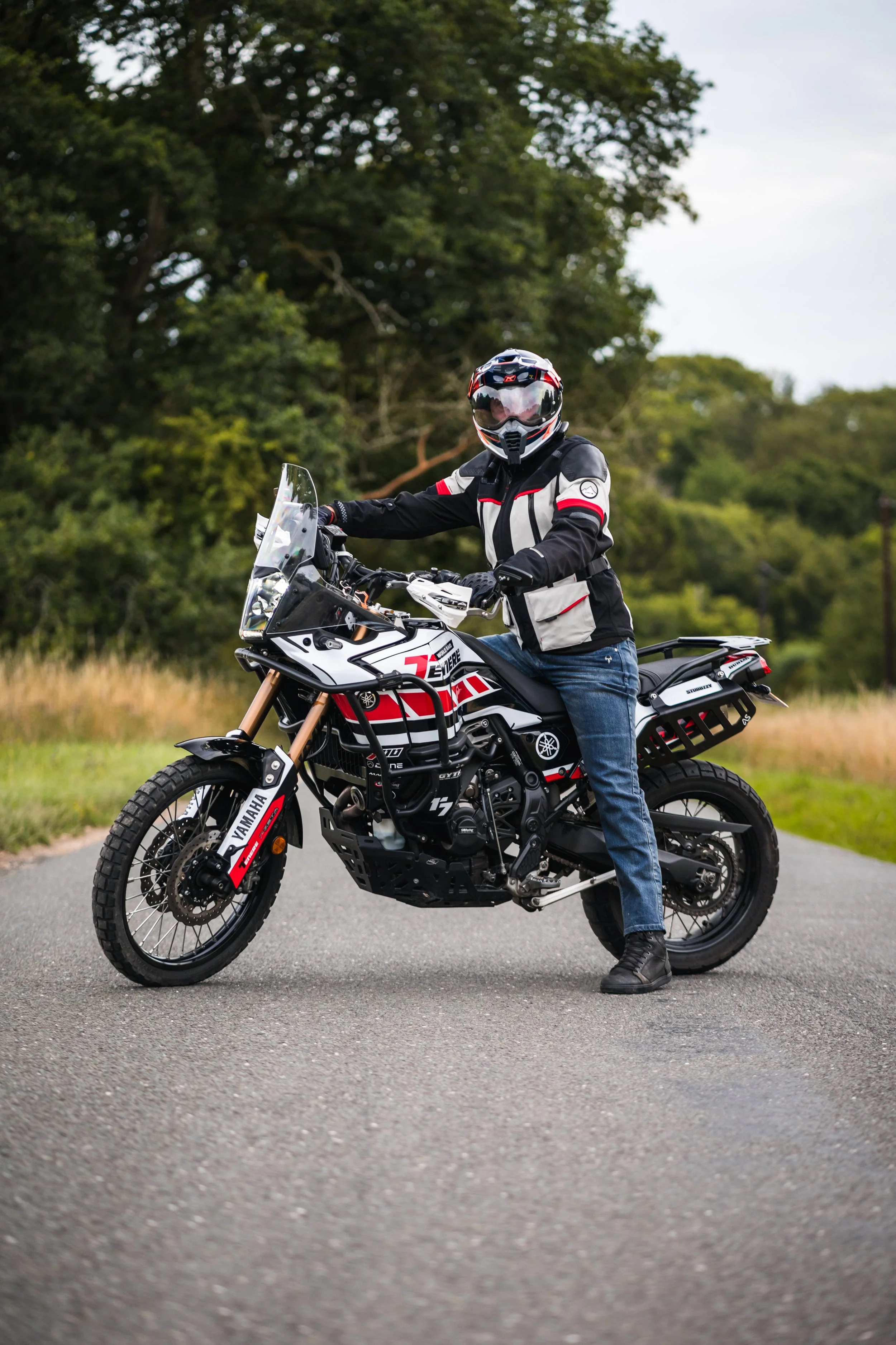 Person wearing motorcycle gear sitting on a Yamaha adventure motorcycle on a paved road with green trees and grass in the background.