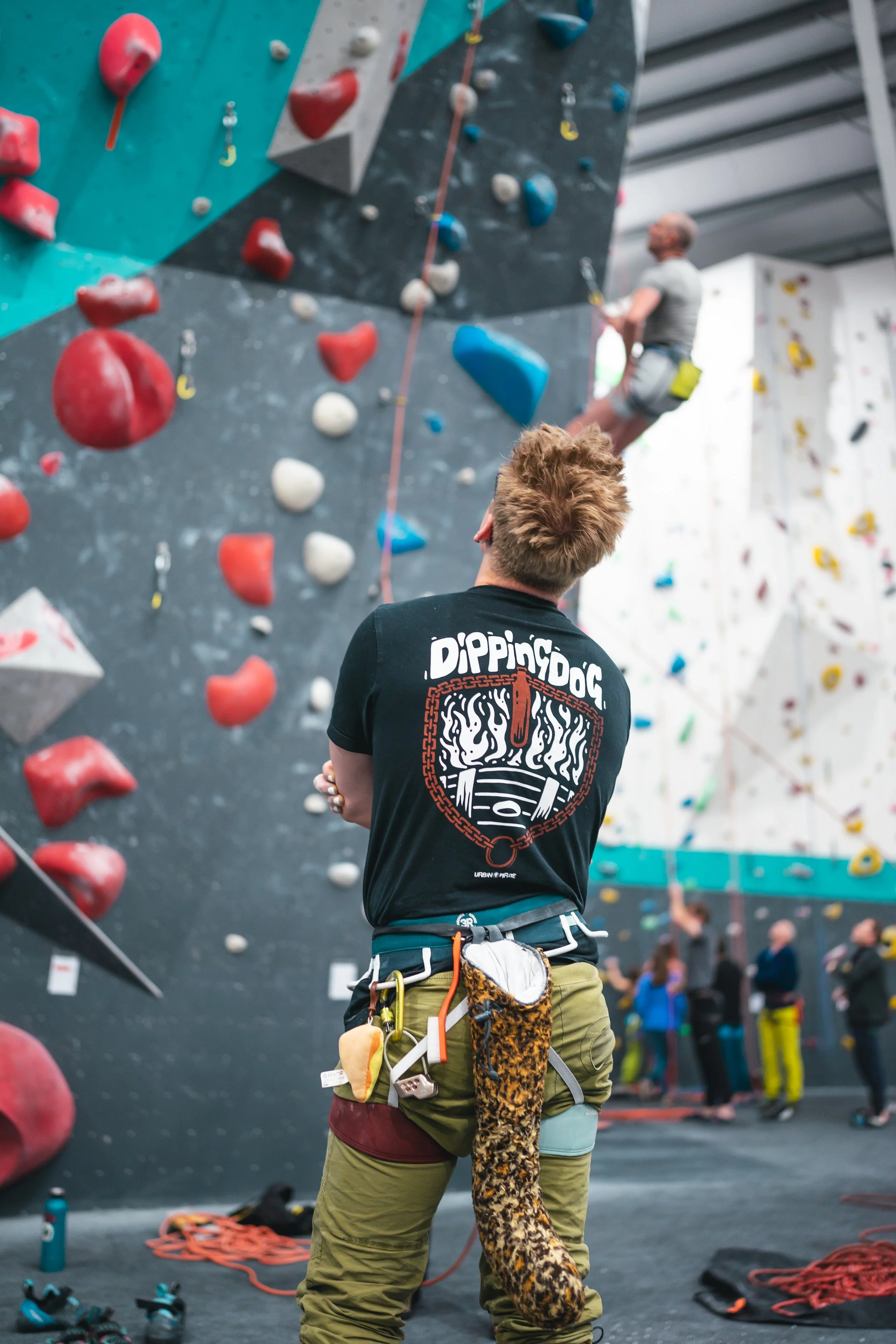 An indoor rock climbing gym with a climber scaling a wall. A belayer watches from below. The wall has colorful holds, and the climber is secured with a harness and rope. Several other people are in the background.