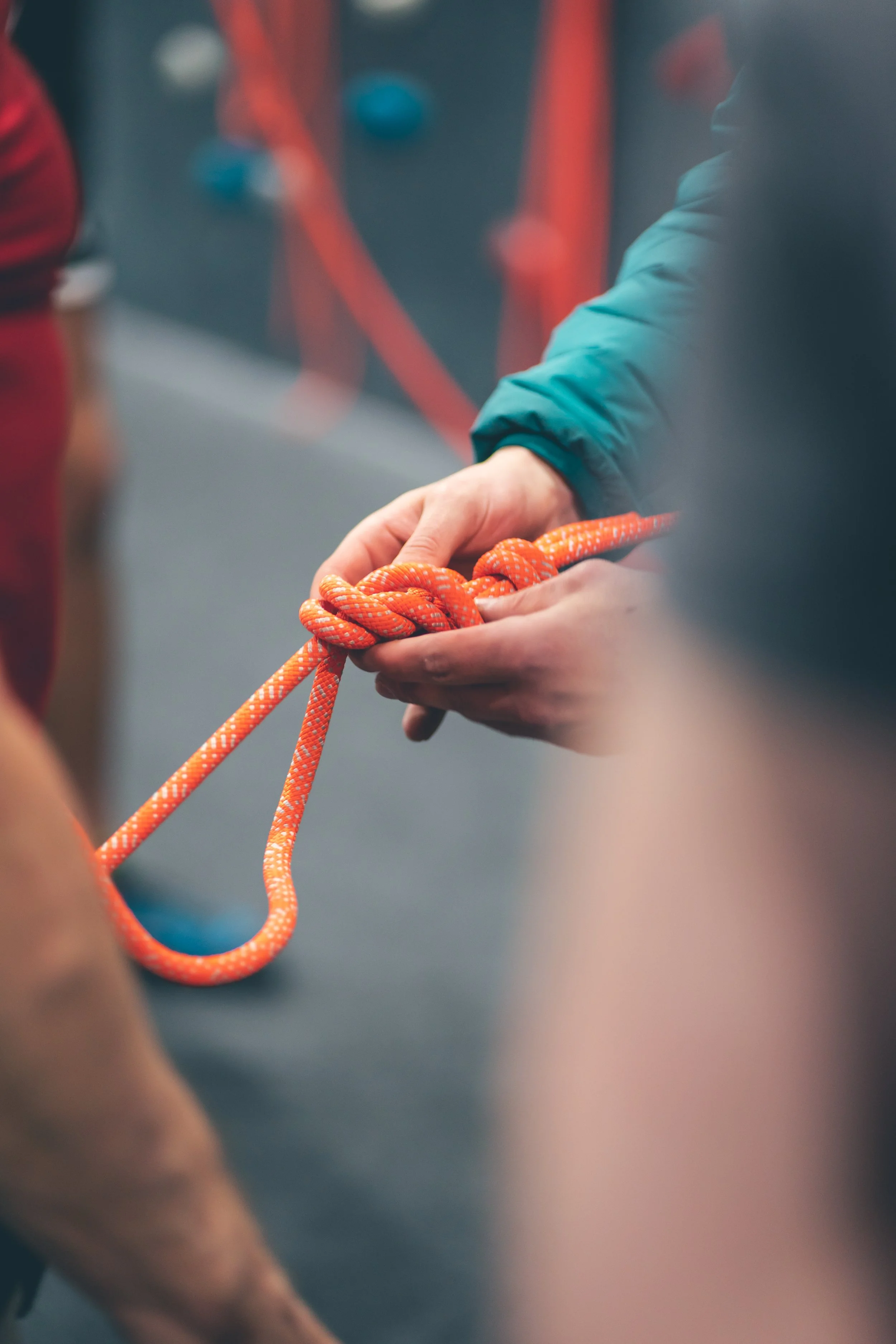 A person in a teal jacket holding a bright orange climbing rope, with others and climbing equipment in the background.