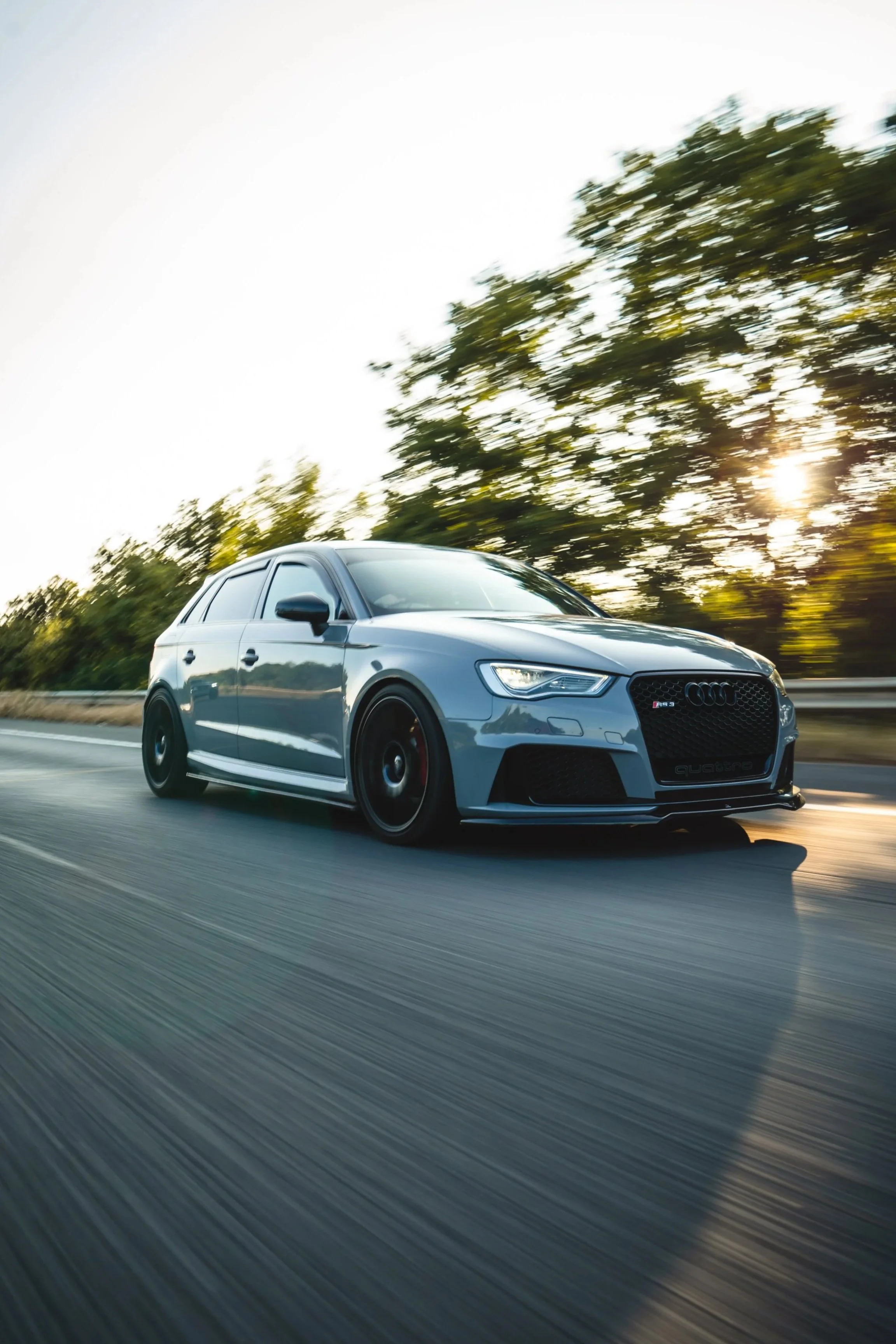 A silver Audi RS3 sedan driving on a highway with trees and a setting sun in the background.