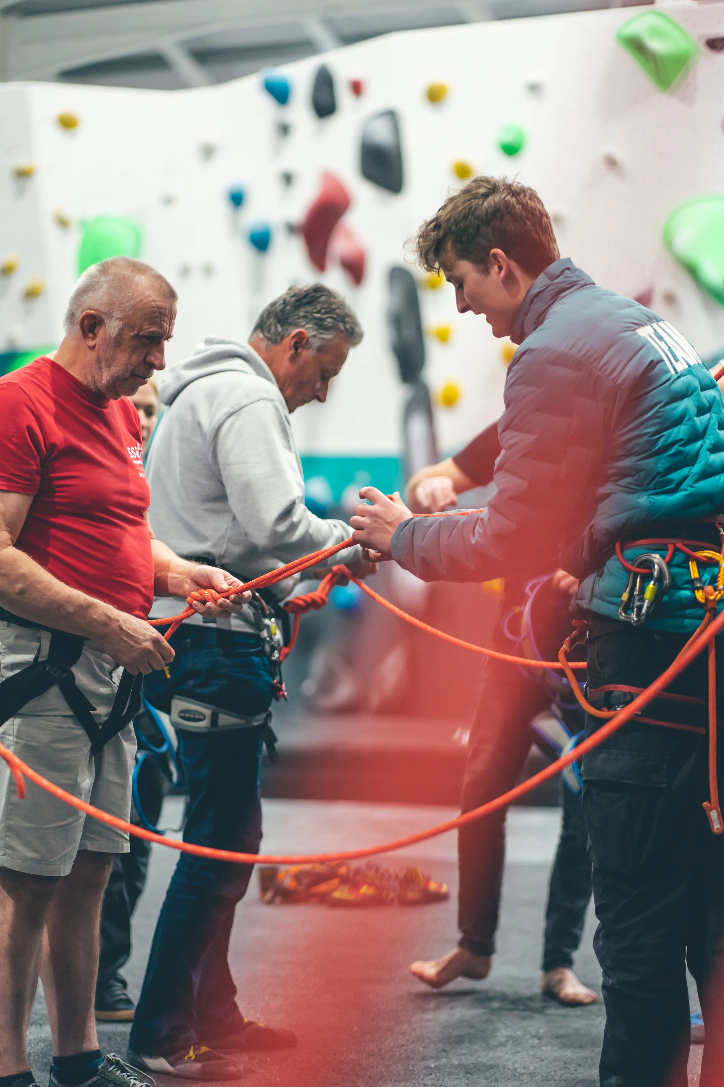 Group of people at an indoor climbing gym, preparing to belay and climb on rock walls with colorful holds.