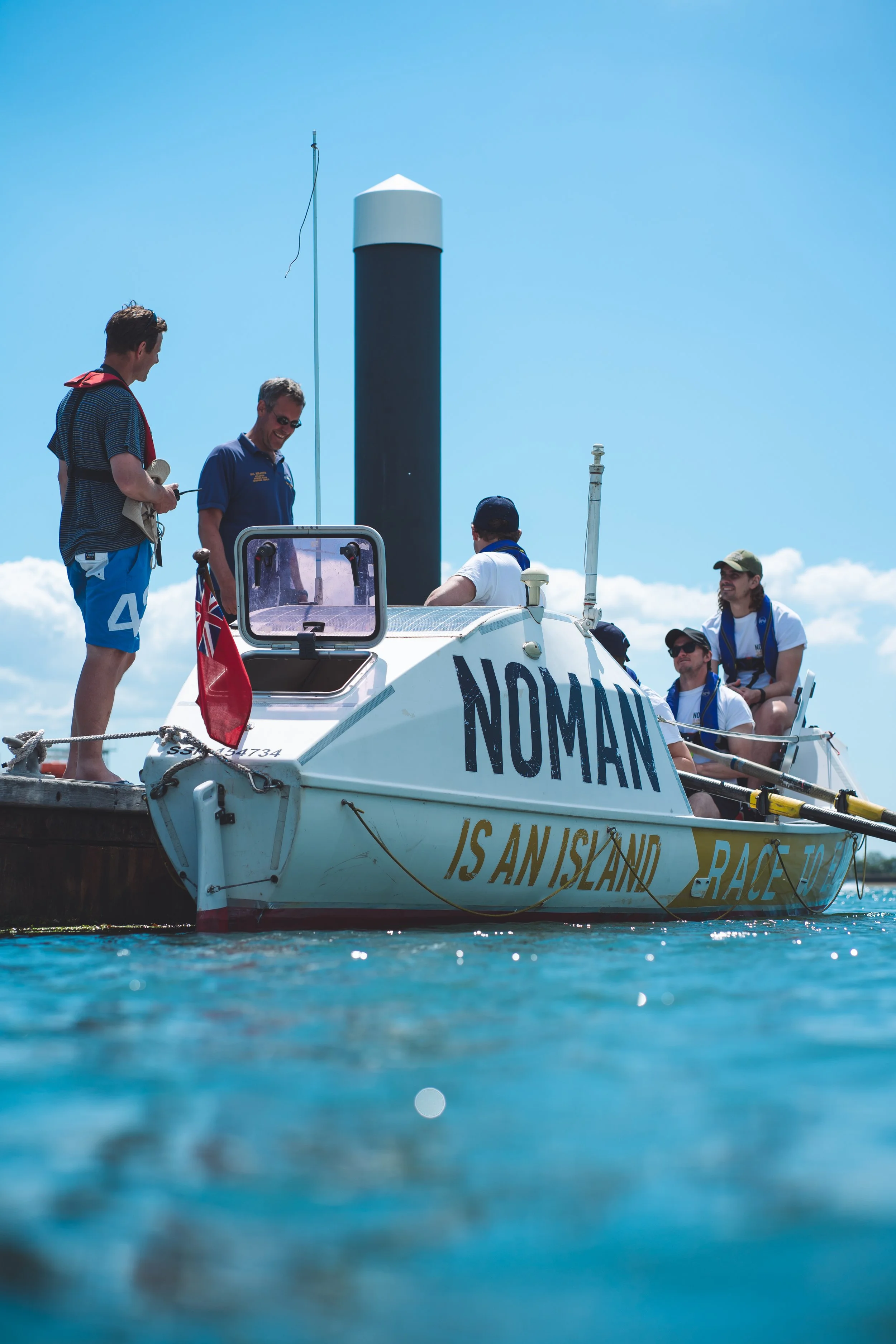 A group of people on a small race boat named 'Nomad' docked at a pier on a sunny day.