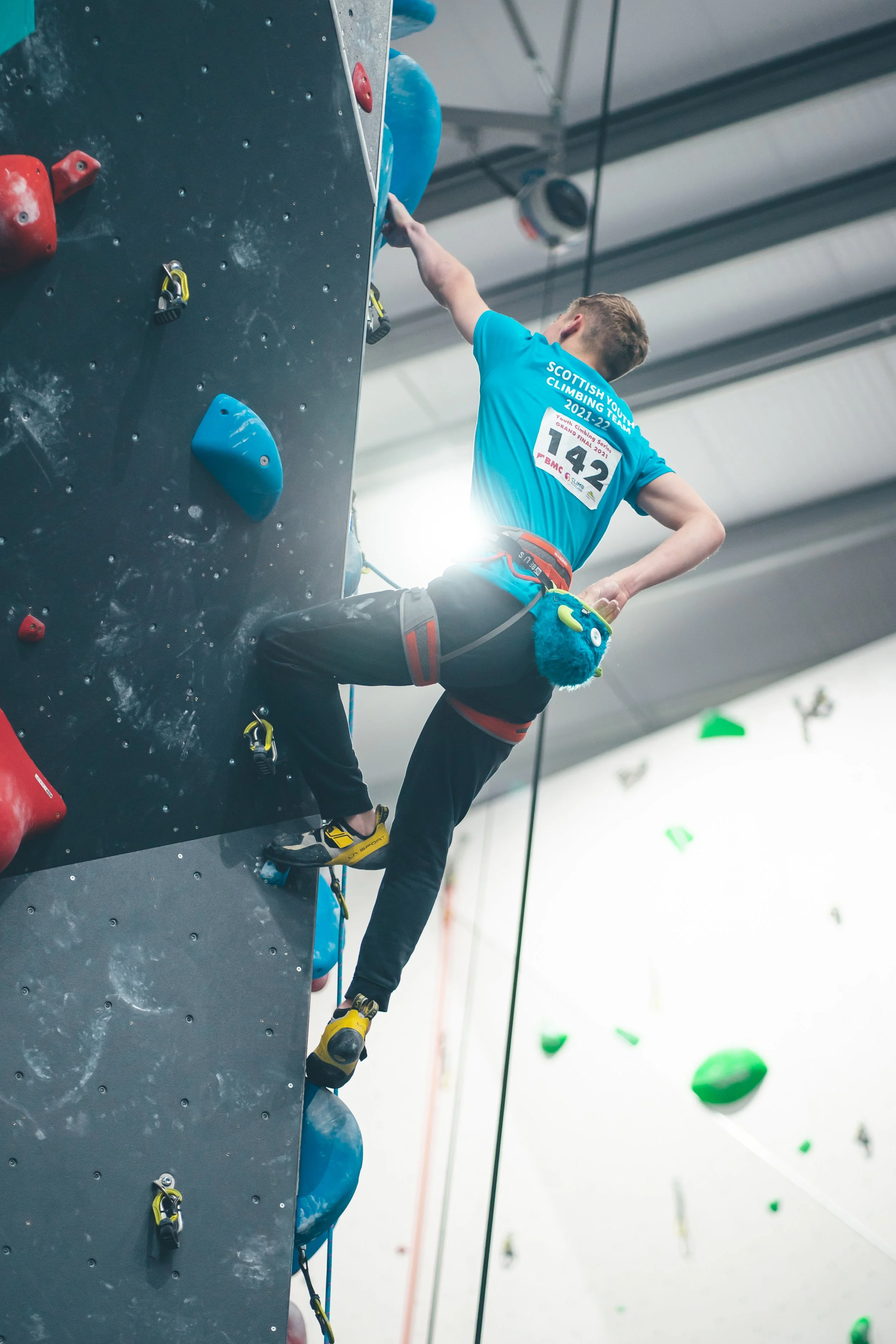 A young male rock climber wearing a blue shirt with a bib number 142 climbs an indoor climbing wall, wearing climbing shoes and a harness.