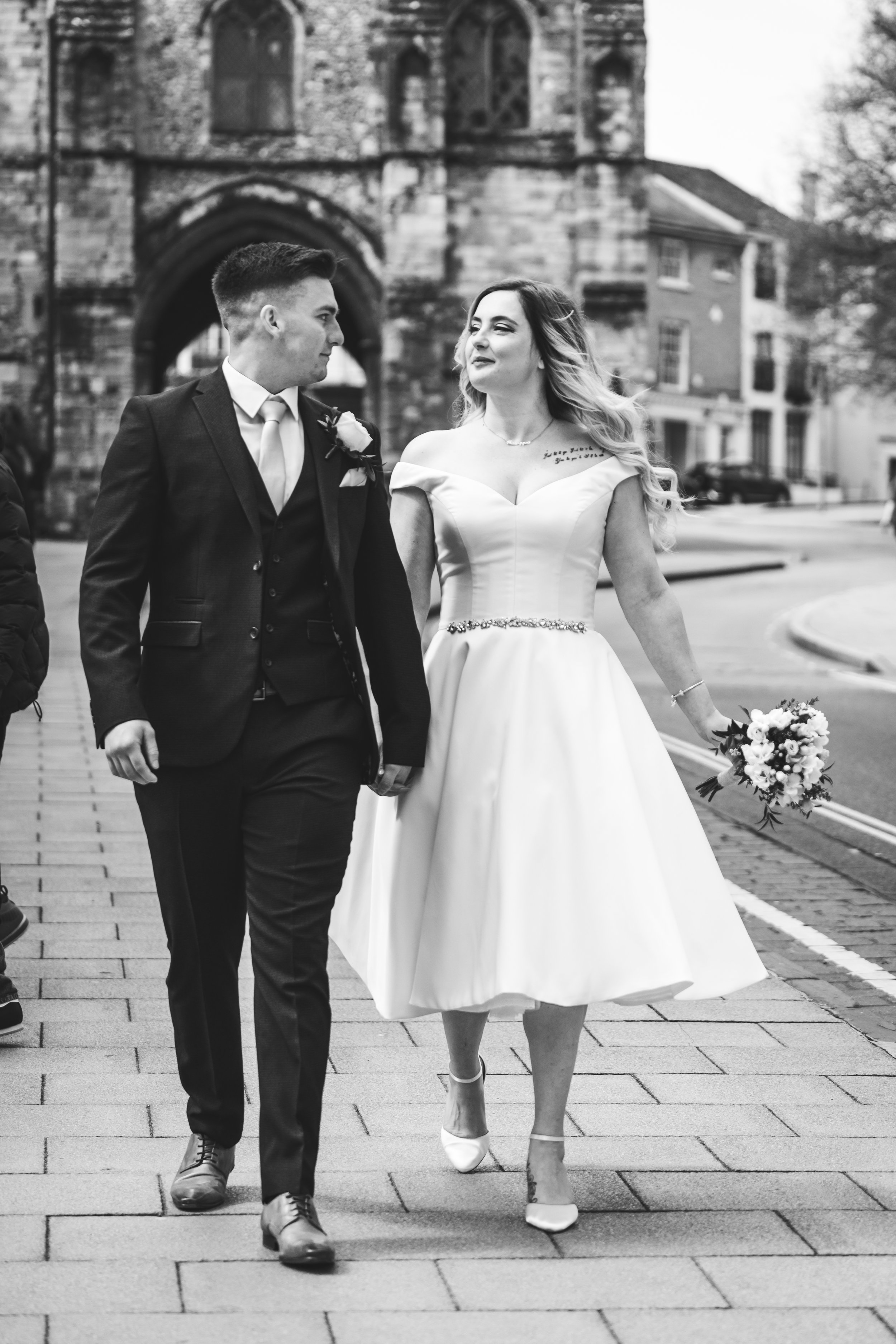 Black and white photo of a bride and groom walking together on a sidewalk in front of an archway and old buildings. The bride is in a vintage-style white dress and holding a bouquet, while the groom is in a dark suit and tie.