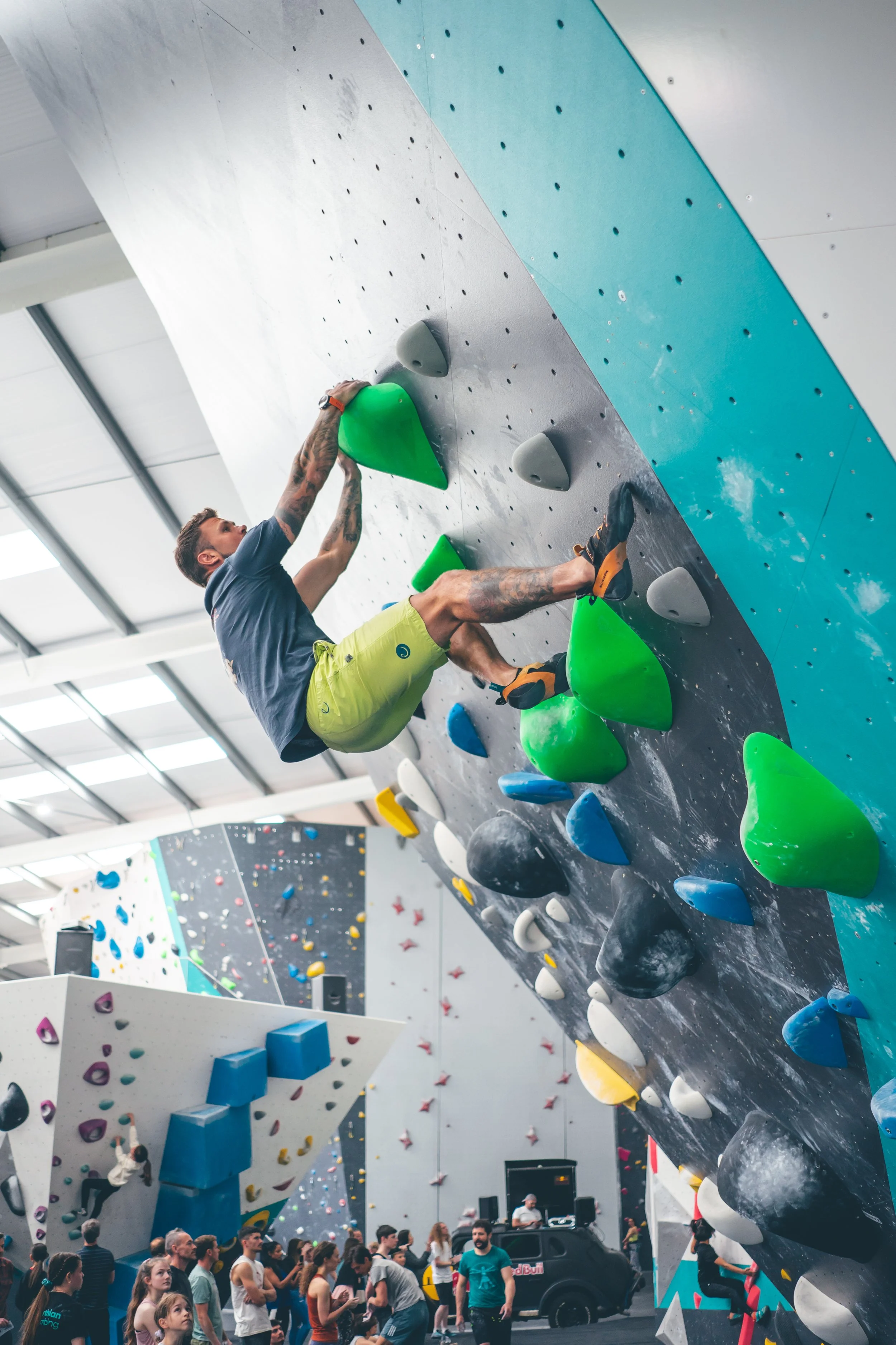 A man bouldering on an indoor climbing wall with various brightly colored holds, while a crowd watches below.