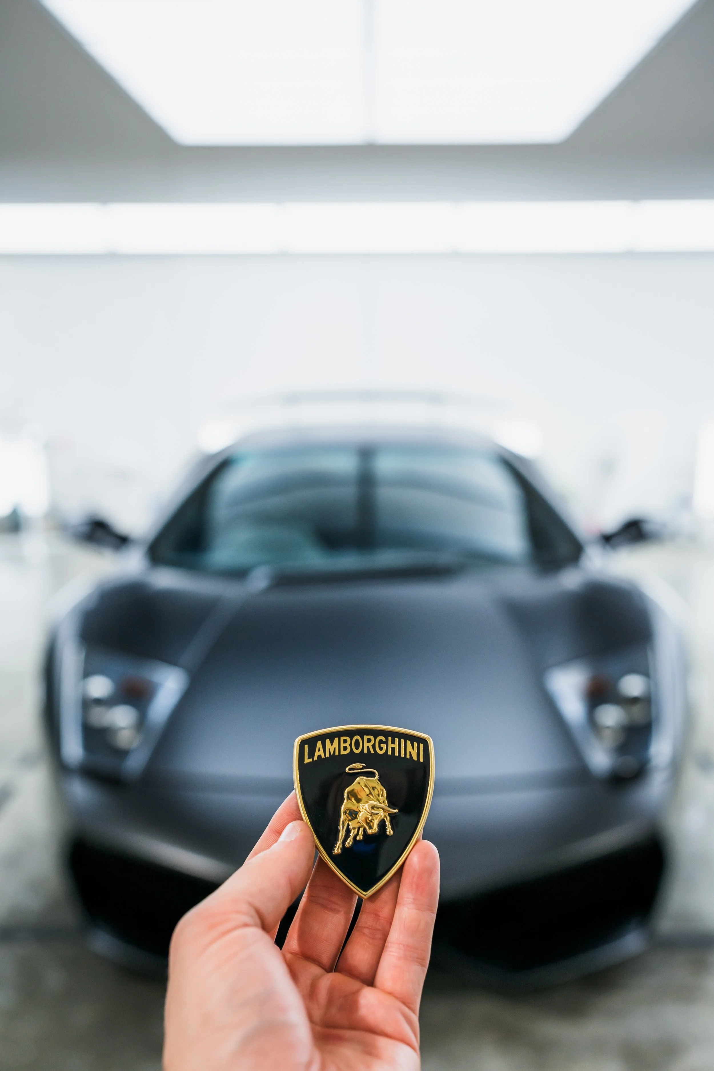 Person holding a Lamborghini badge in front of a sleek black Lamborghini sports car inside a showroom.