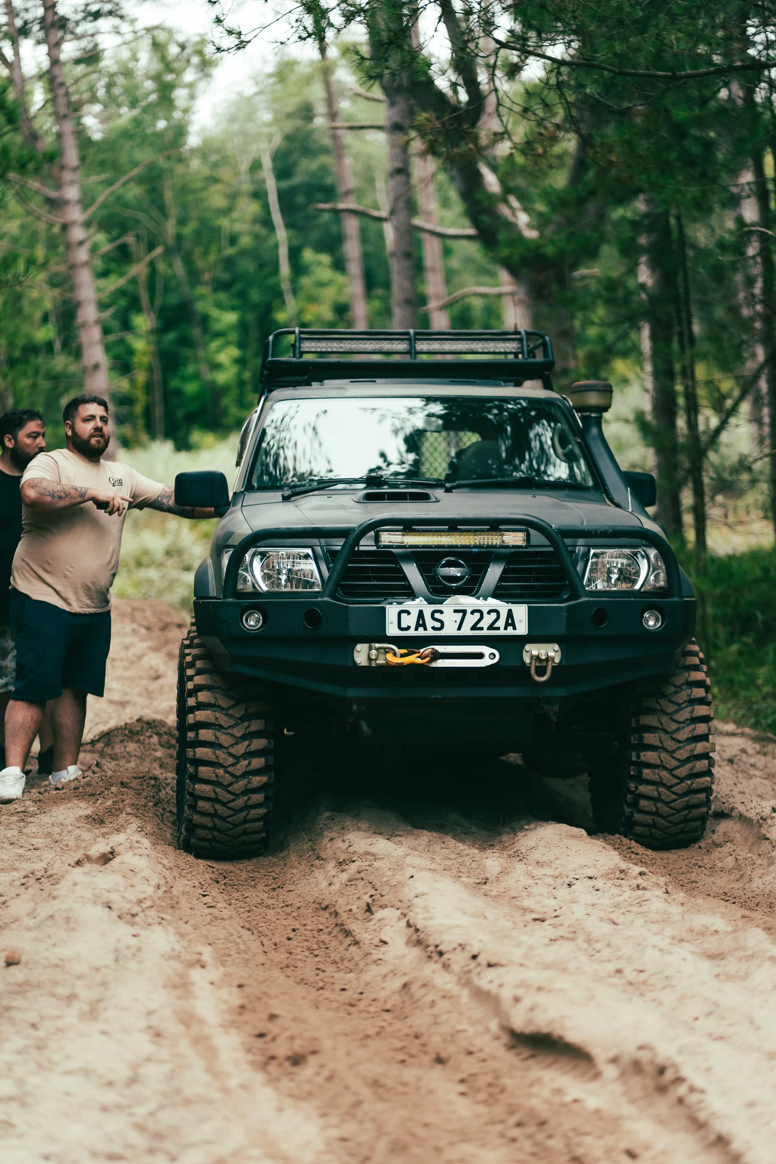 A black off-road vehicle with large tires and a front bumper winch, stuck in sandy terrain in a forest, with two men pulling on the vehicle's side mirror.