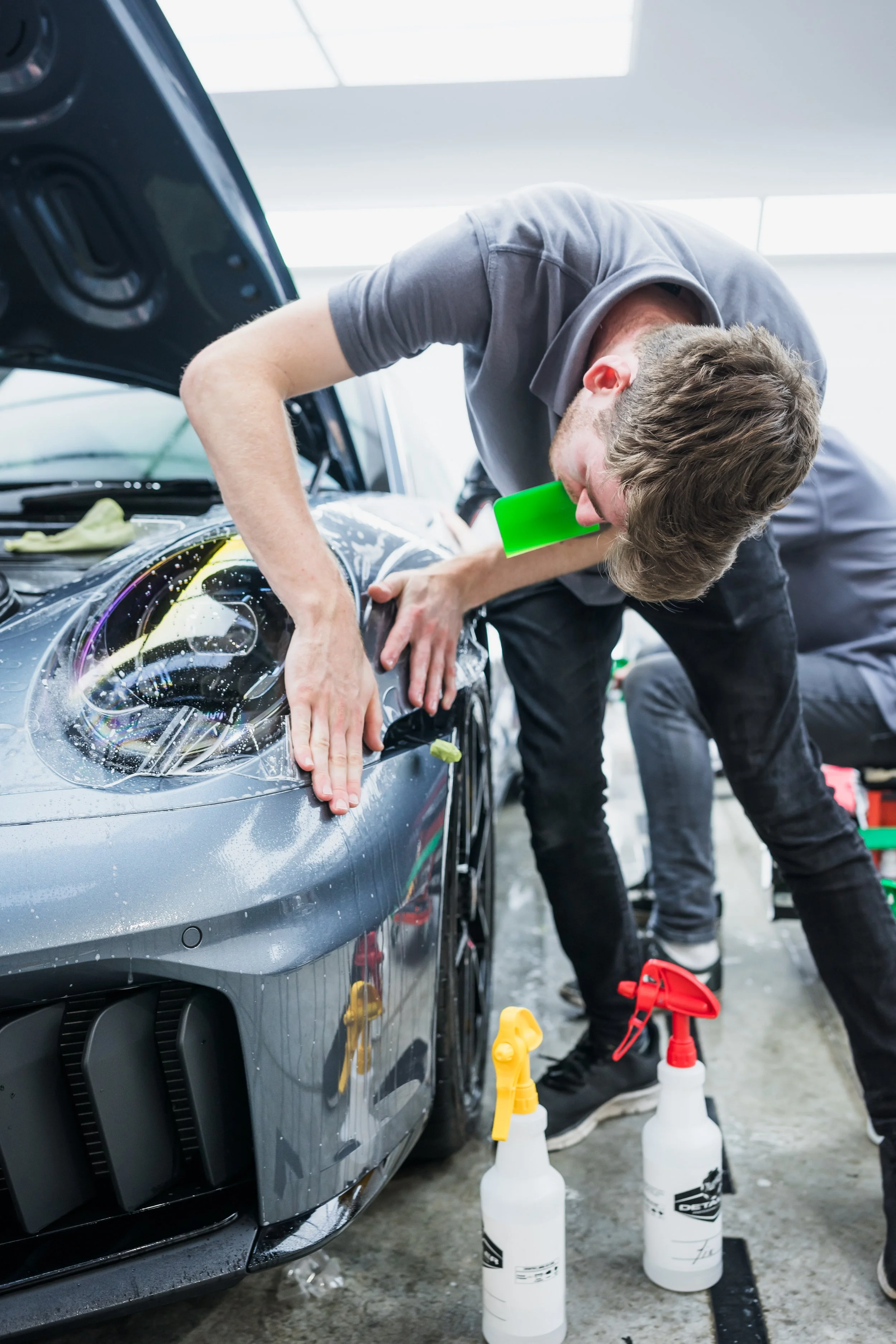 A man cleaning a gray sports car with soap and a green sponge in an auto detail shop.