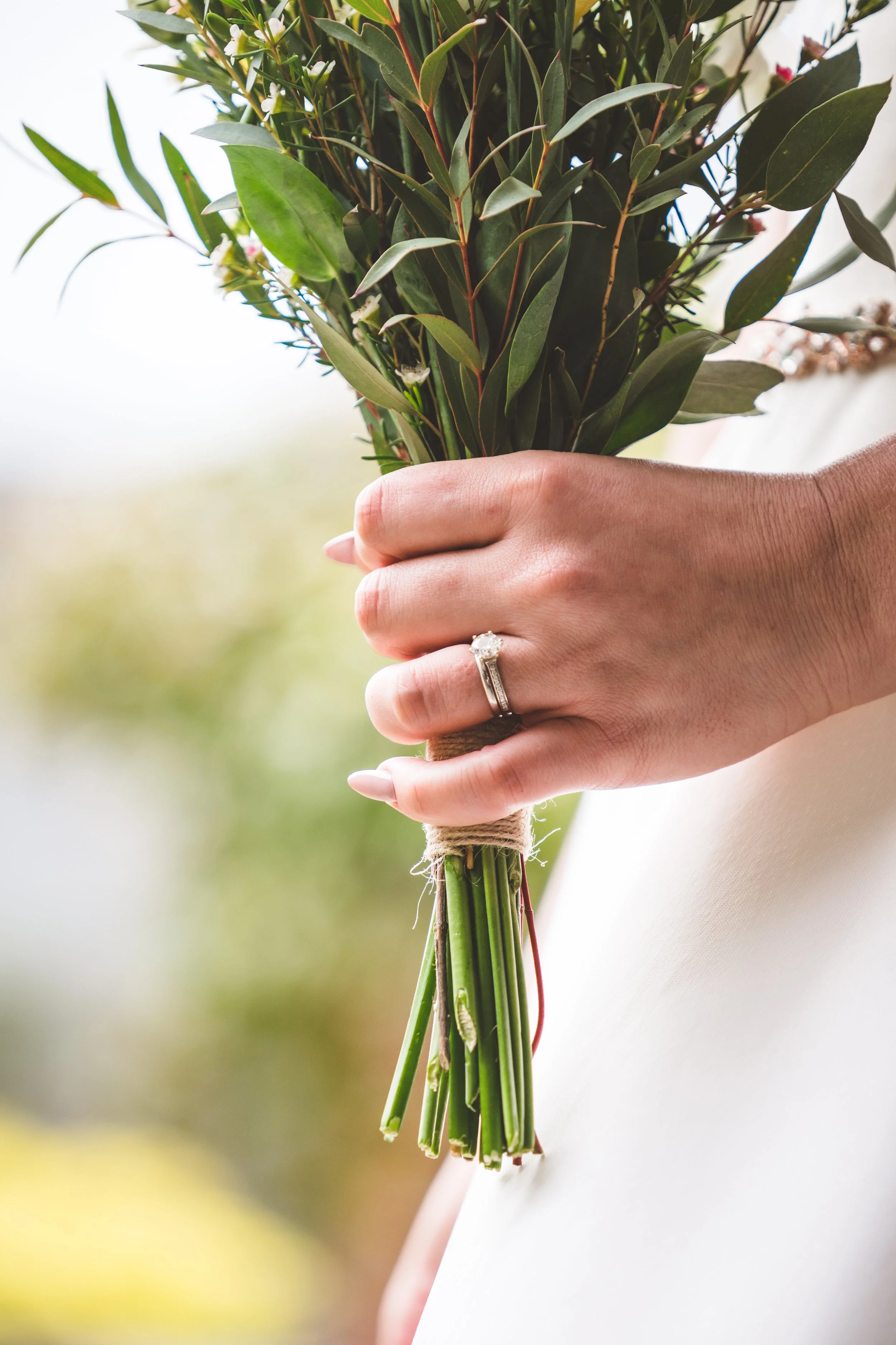 A woman holding a bouquet of green foliage and pink flowers, with a wedding ring on her finger.