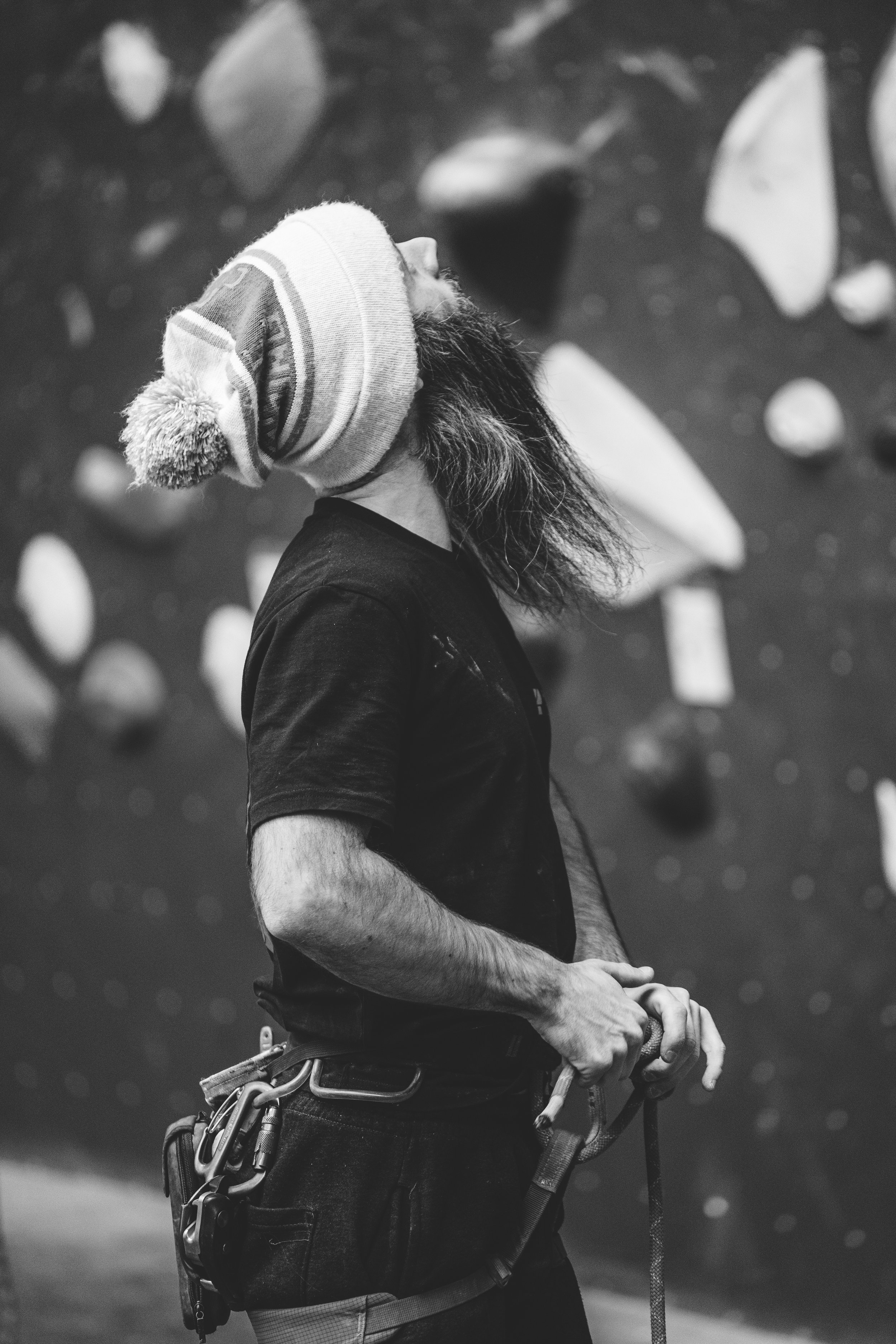 A man with long hair wearing a knit beanie hat, black t-shirt, and climbing harness stands against a background of a rock climbing wall.