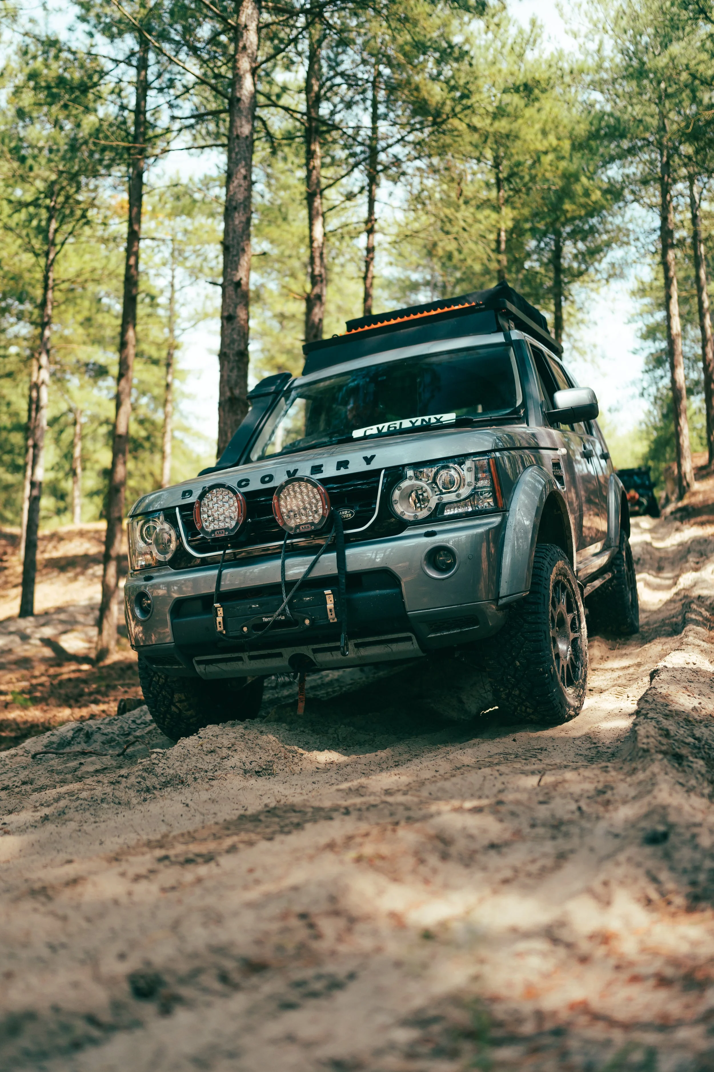 A grey off-road vehicle with front-mounted lights and a roof box navigating a sandy trail in a forest with tall pine trees.