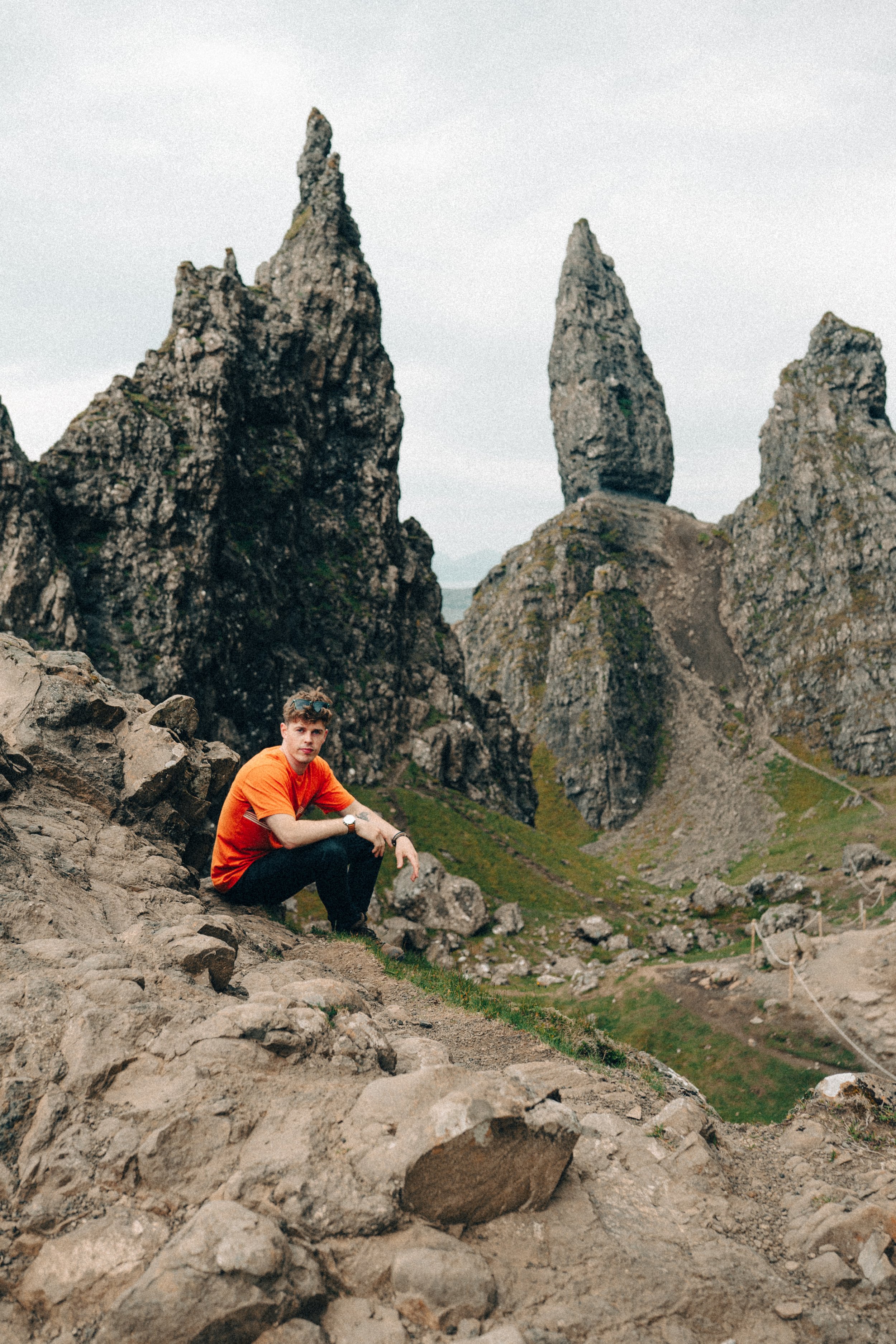 A young man in an orange shirt crouching on rocky terrain with towering jagged rock formations in the background, under an overcast sky.