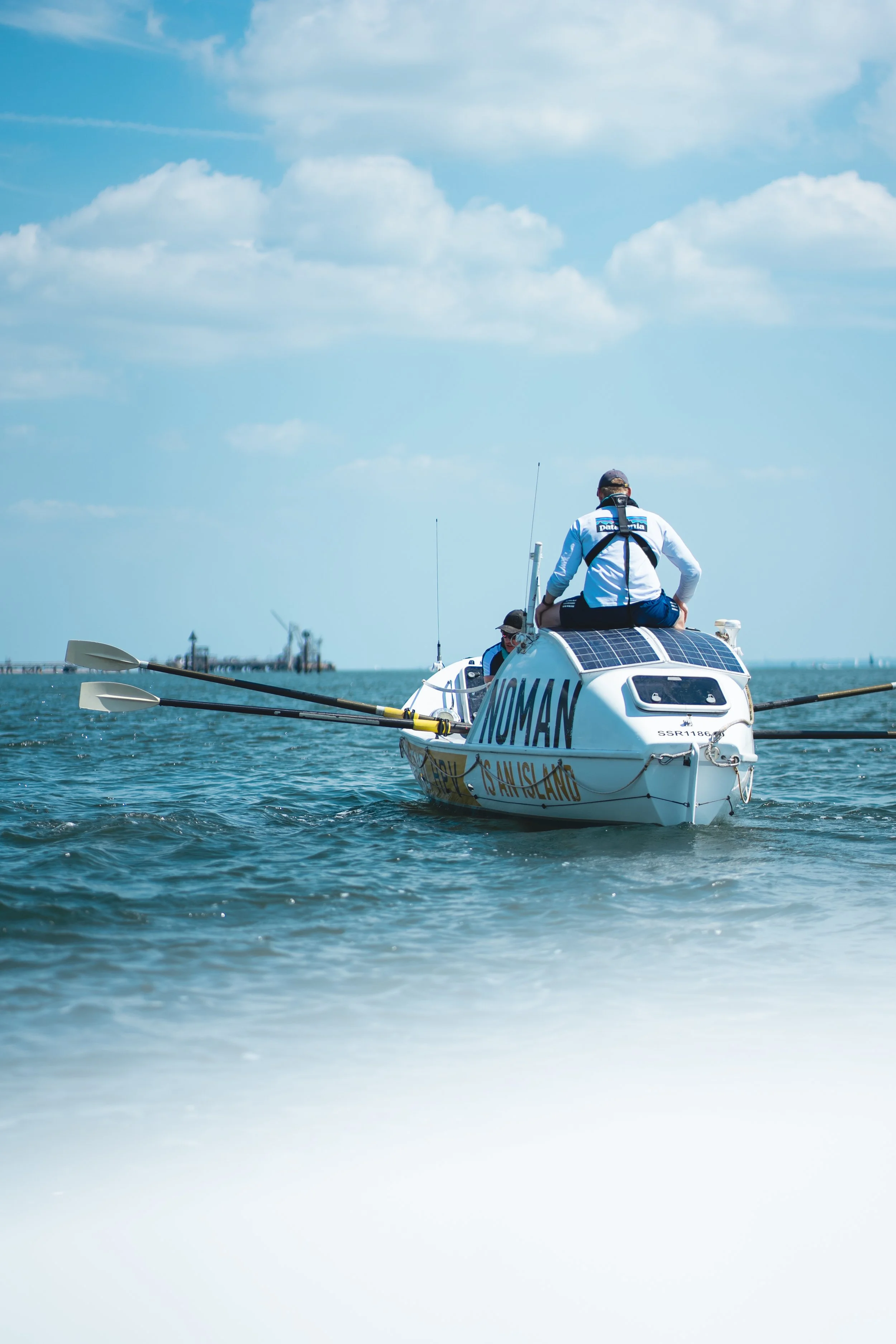 A person in a white jacket with blue accents sitting on the back of a small racing boat with the word 'NOMAN' on it, on the water with a clear blue sky and clouds.
