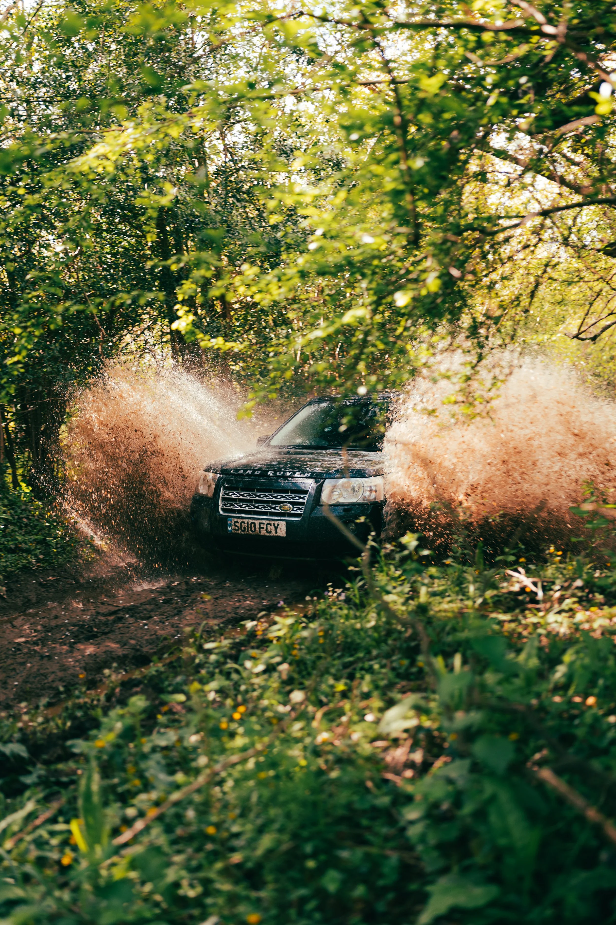 A black Land Rover driving through a muddy forest trail, splashing water and mud, surrounded by green foliage and trees.