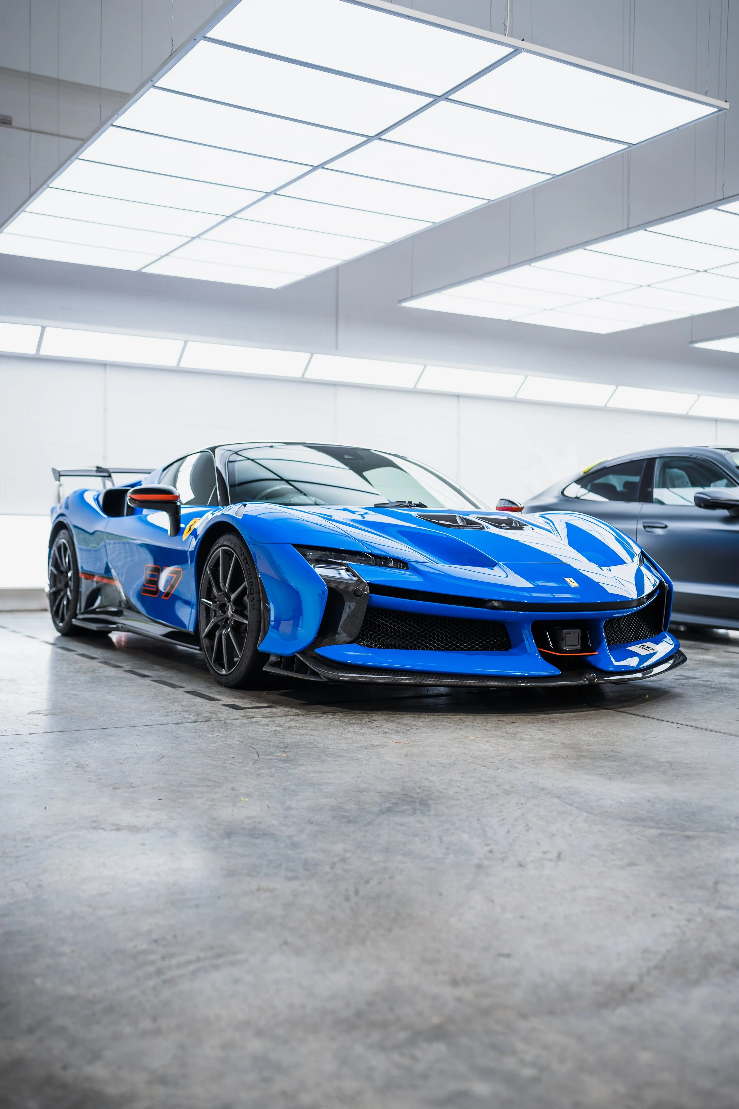 A blue race car with black accents parked indoors, next to a gray vehicle, under bright white ceiling lights.