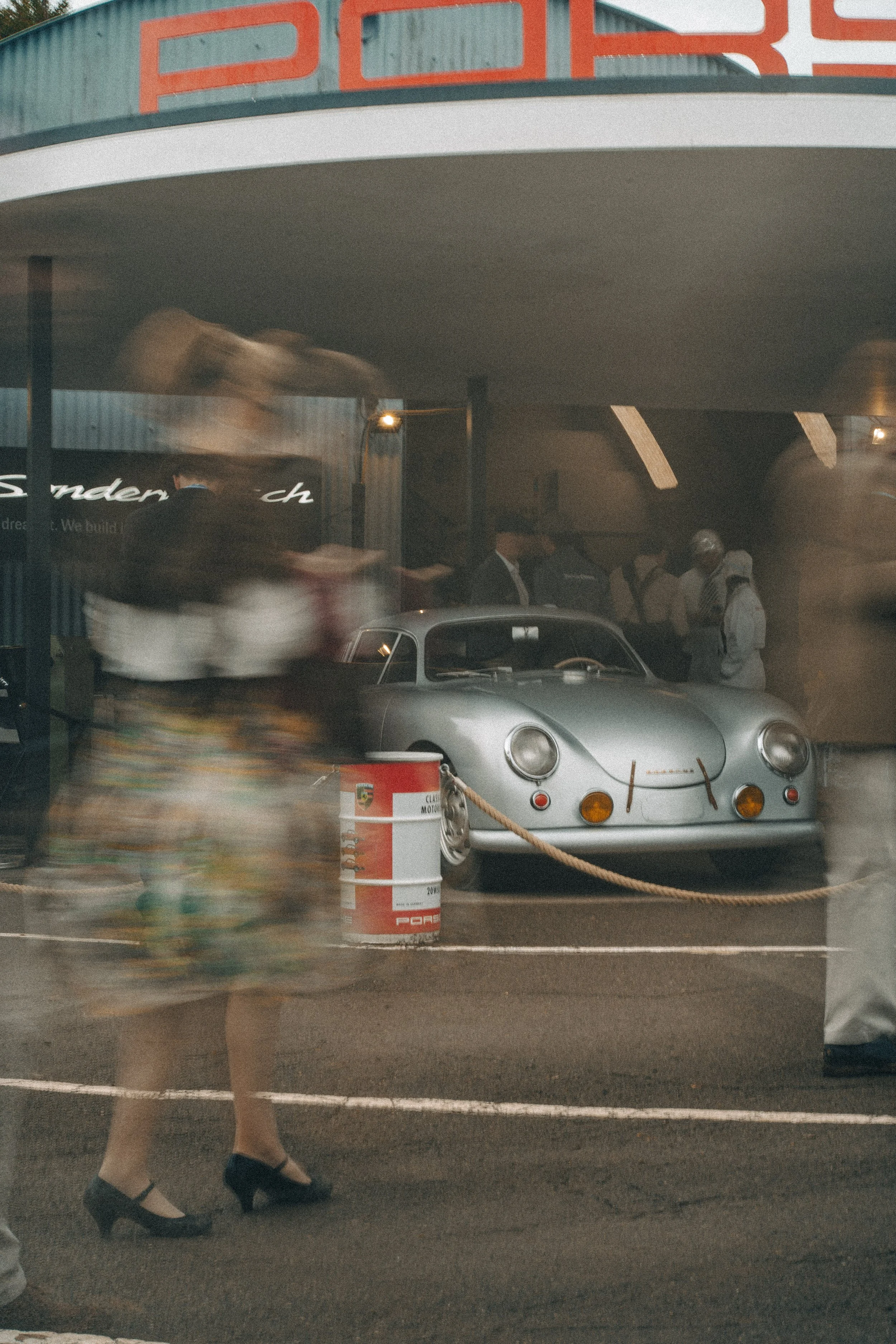 A classic silver Porsche sports car on display behind a rope barrier at an exhibition, with a blurred woman in colorful clothing walking past in the foreground, and a group of people in the background under a building with a large red sign.