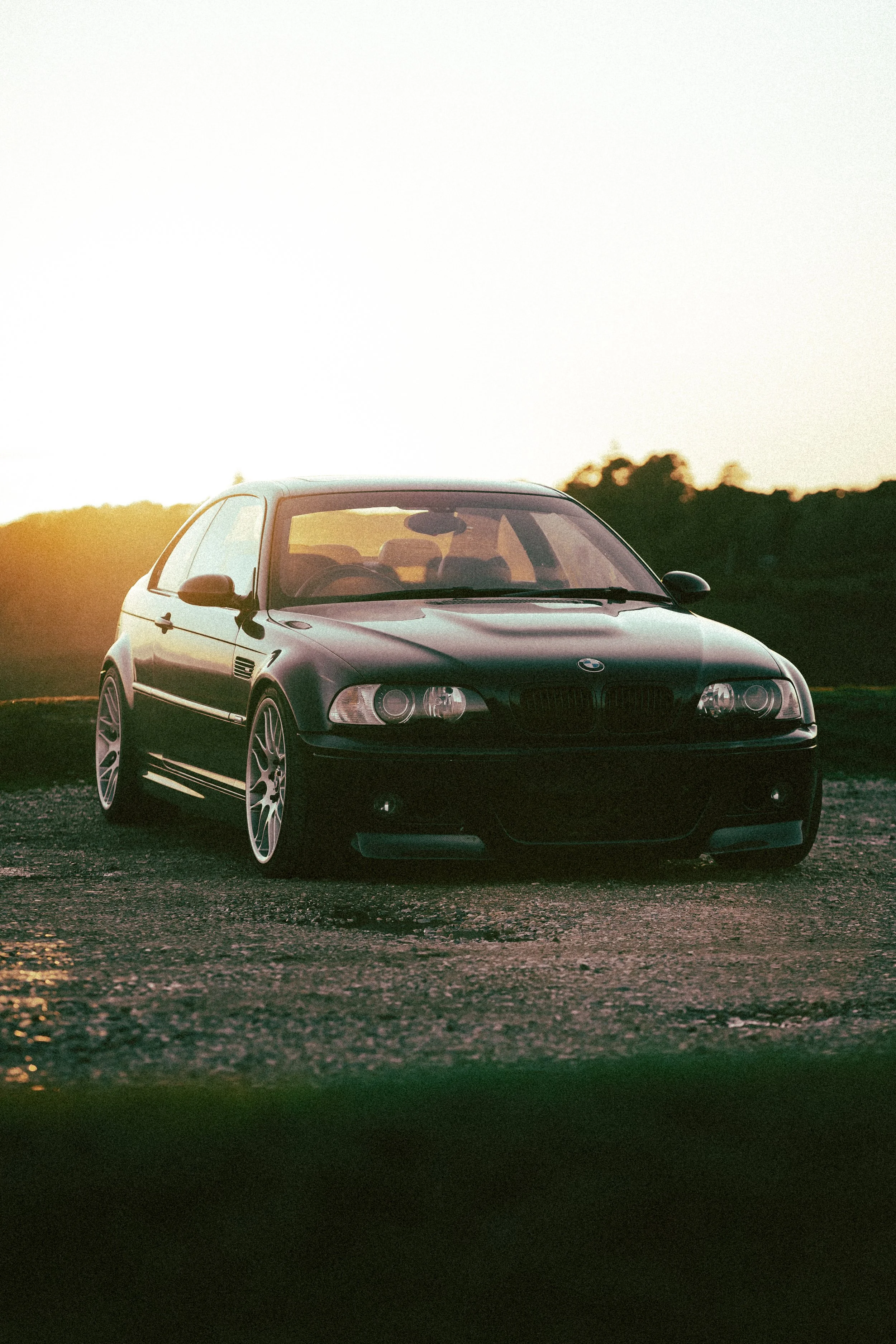 A black BMW car parked on a gravel surface at sunset, with trees in the background.