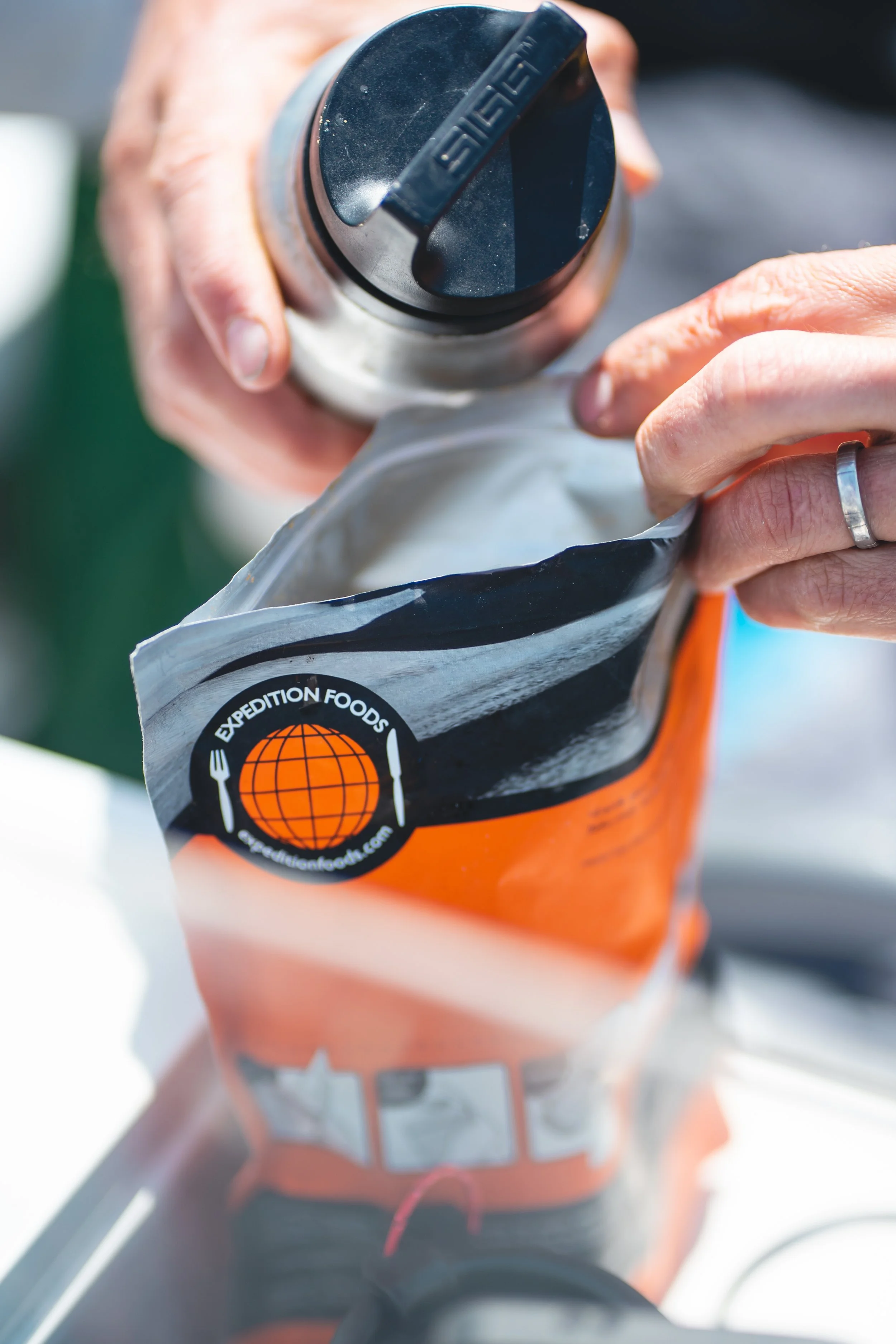 A close-up of hands opening a bag of Expedition Foods emergency food, with a person's hand holding a black-lidded shaker bottle over the bag.