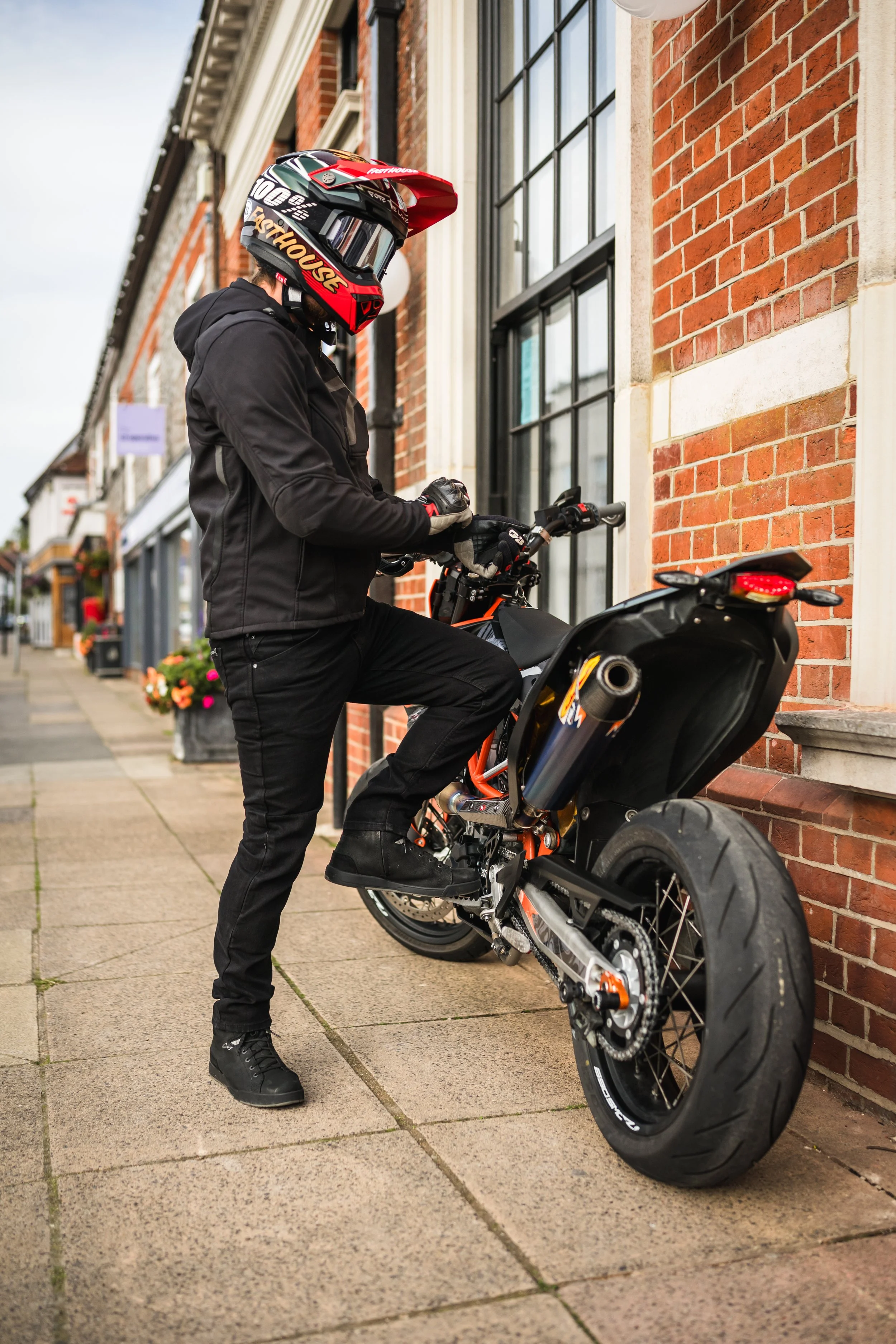 Motorcyclist wearing a helmet, gloves, and black clothing, standing beside a black and orange motorcycle parked against a brick building on the sidewalk.