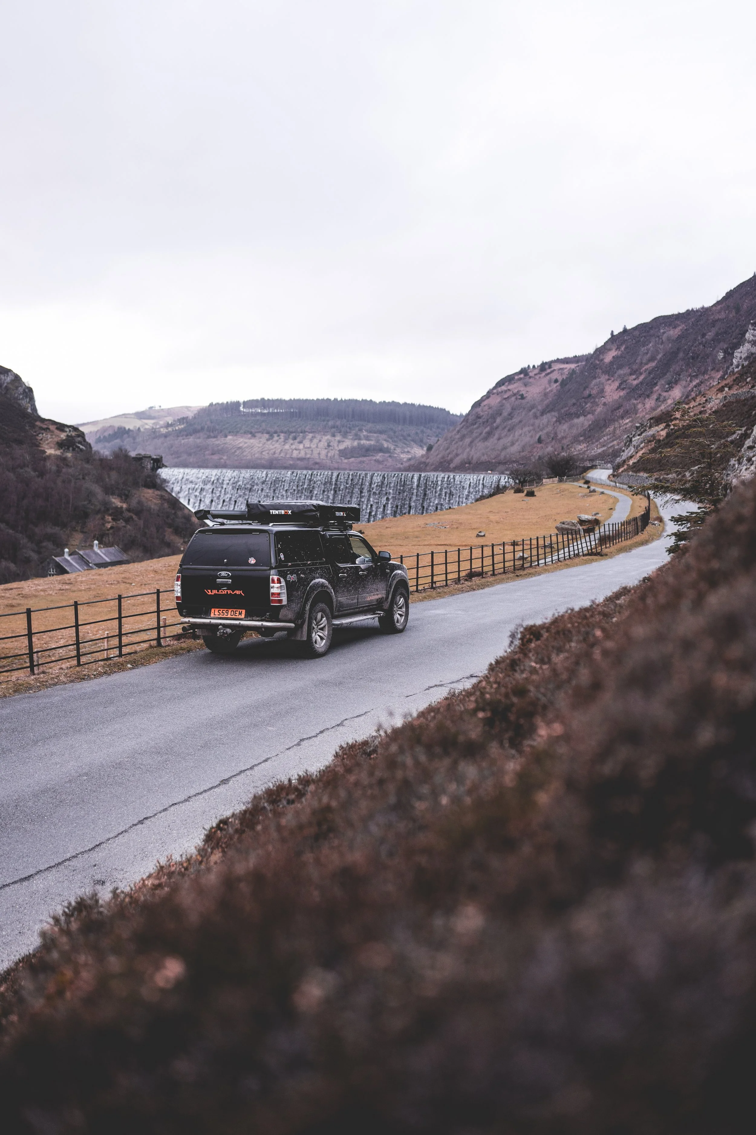 A black SUV driving on a winding road through a hilly landscape with a dam in the background, overcast sky.