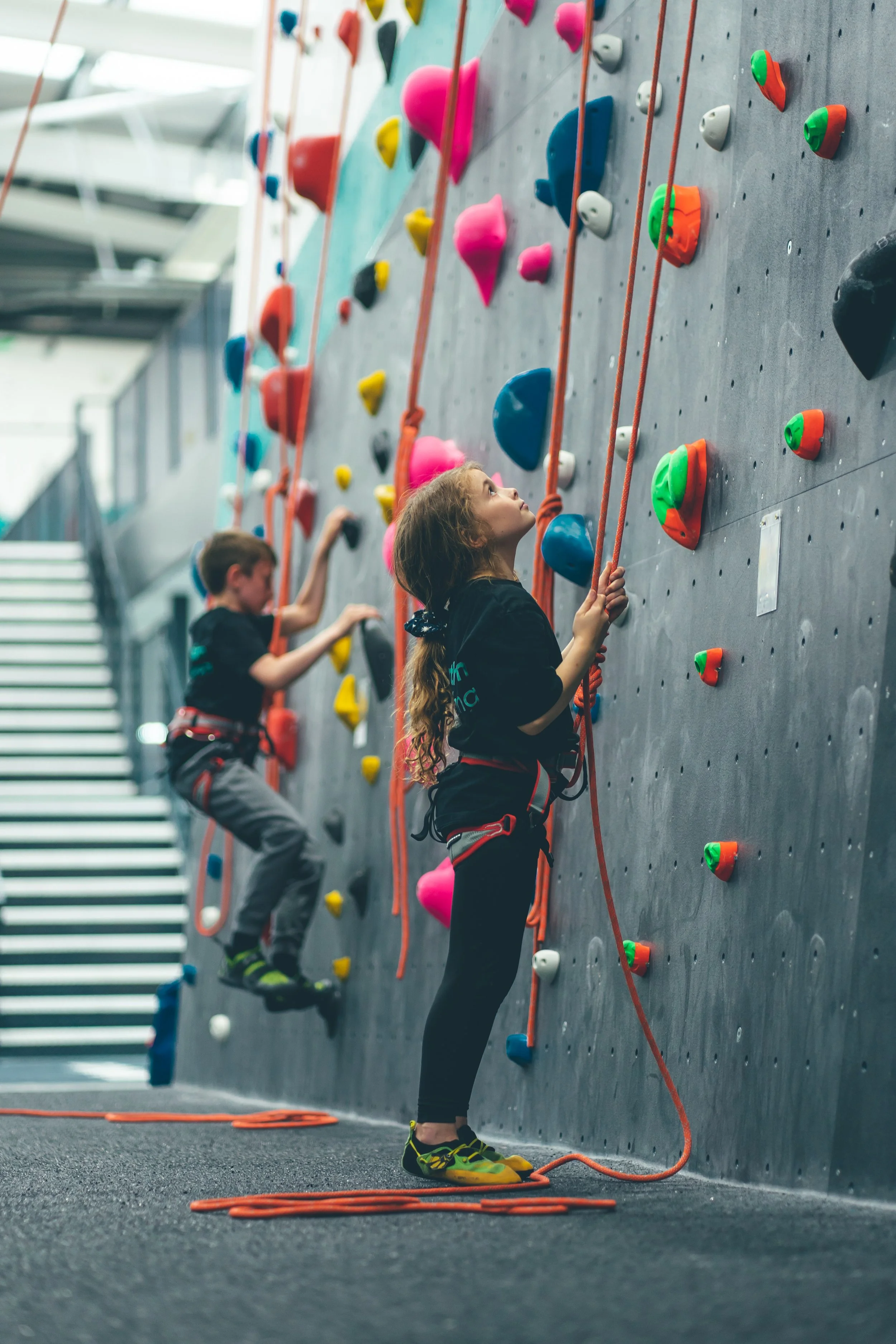 Two children, a boy and a girl, climbing an indoor rock wall with colorful holds and orange ropes, in a gym setting.