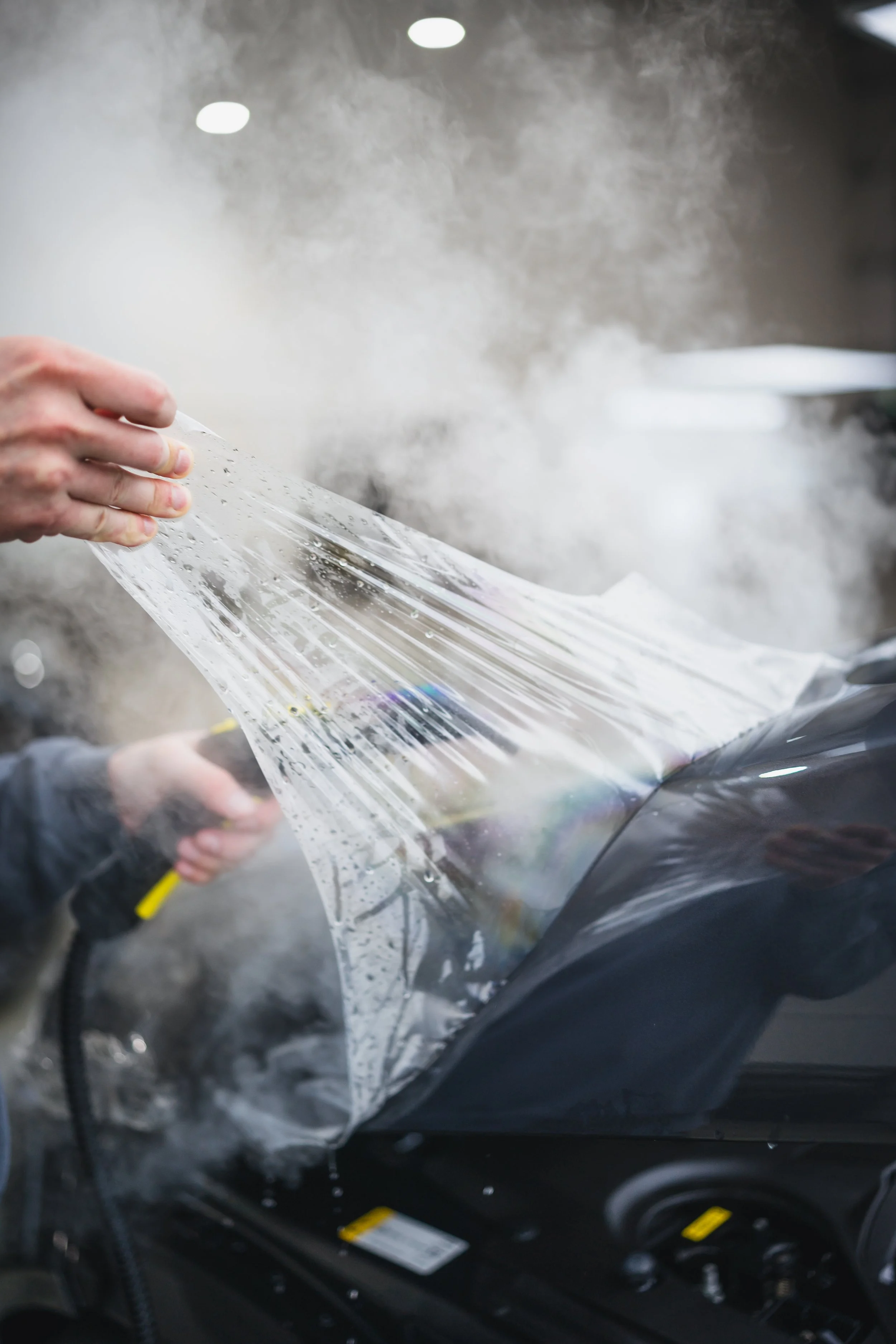 A person removing a protective plastic film from a car's hood, with visible steam or vapor surrounding the area.