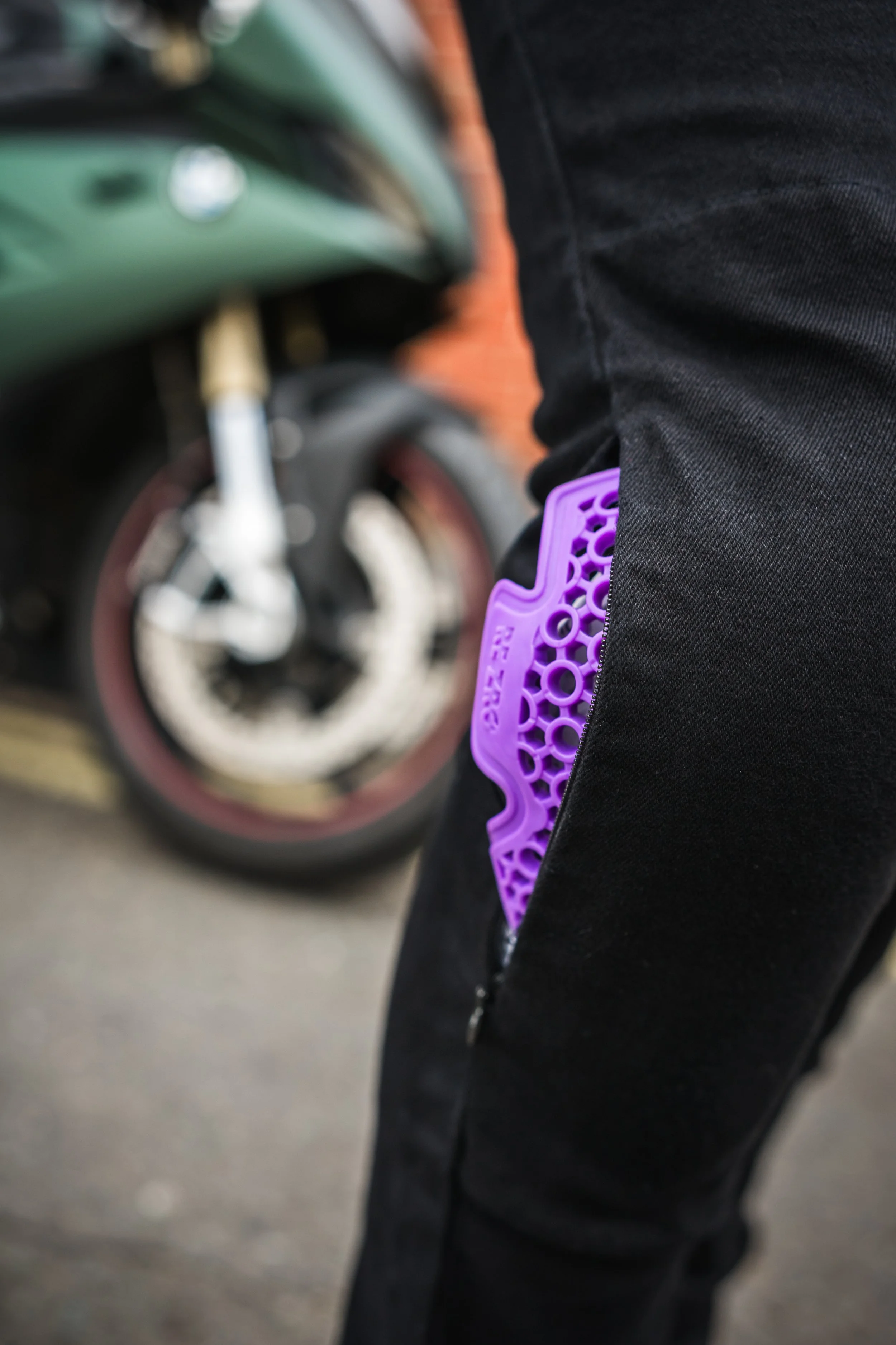 A person wearing black pants with a purple item, possibly a grip or grip cover, protruding from their pocket, standing beside a motorcycle with a green body and a disc brake, on a paved surface with a brick wall in the background.
