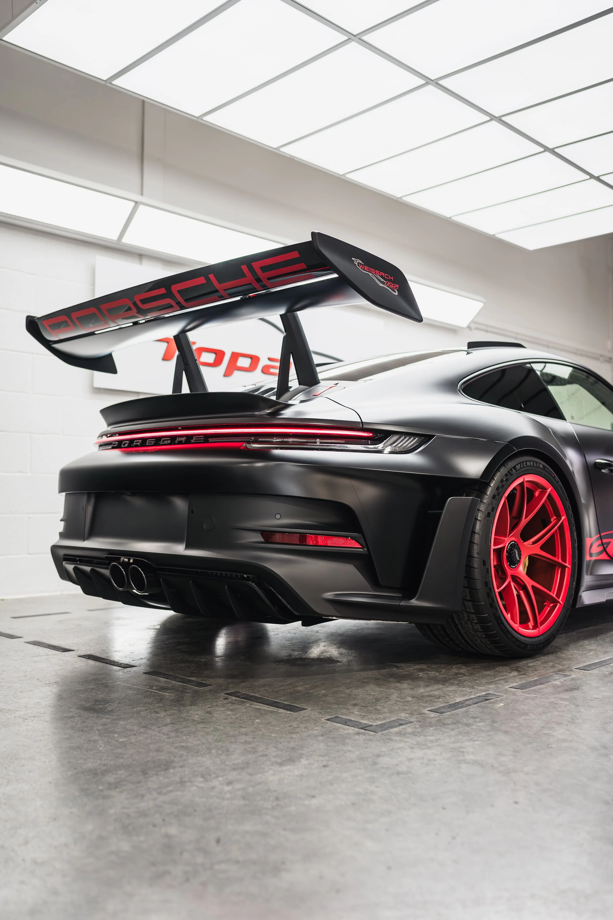 Rear view of black Porsche 911 with red accents, racing wing, dual exhaust, and distinctive red wheels in a garage.