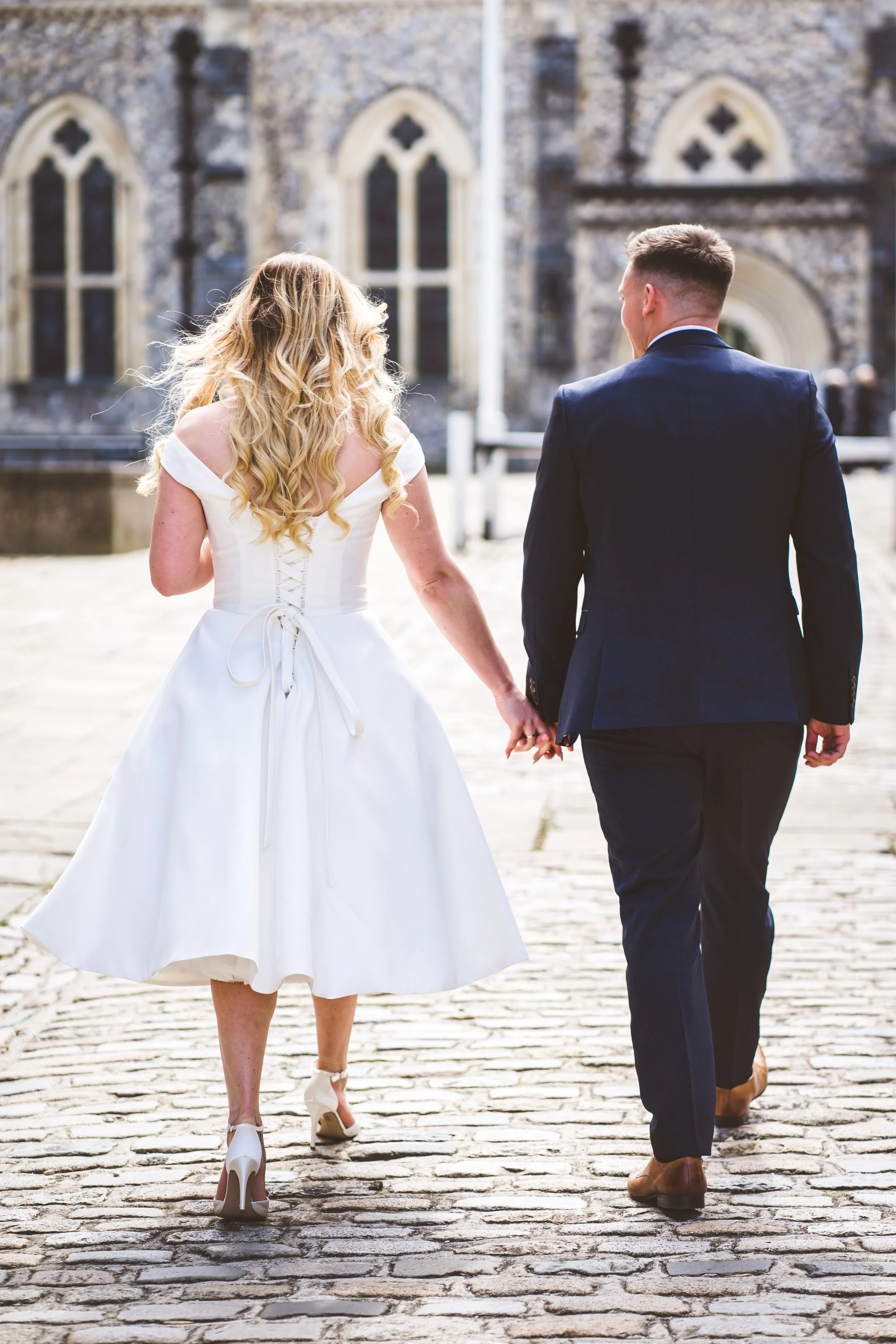 A bride and groom walking hand in hand outside a historic church, seen from behind, with the bride wearing a white wedding dress and the groom in a dark suit.