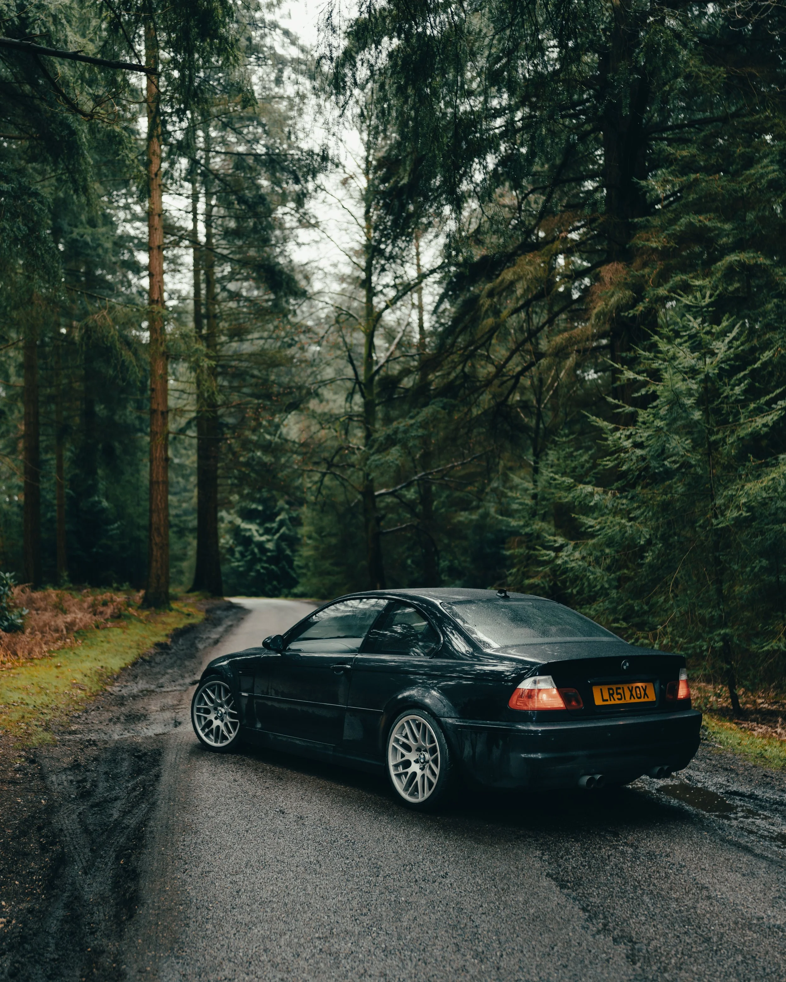 Black convertible car parked on a wet, winding forest road surrounded by tall green trees.