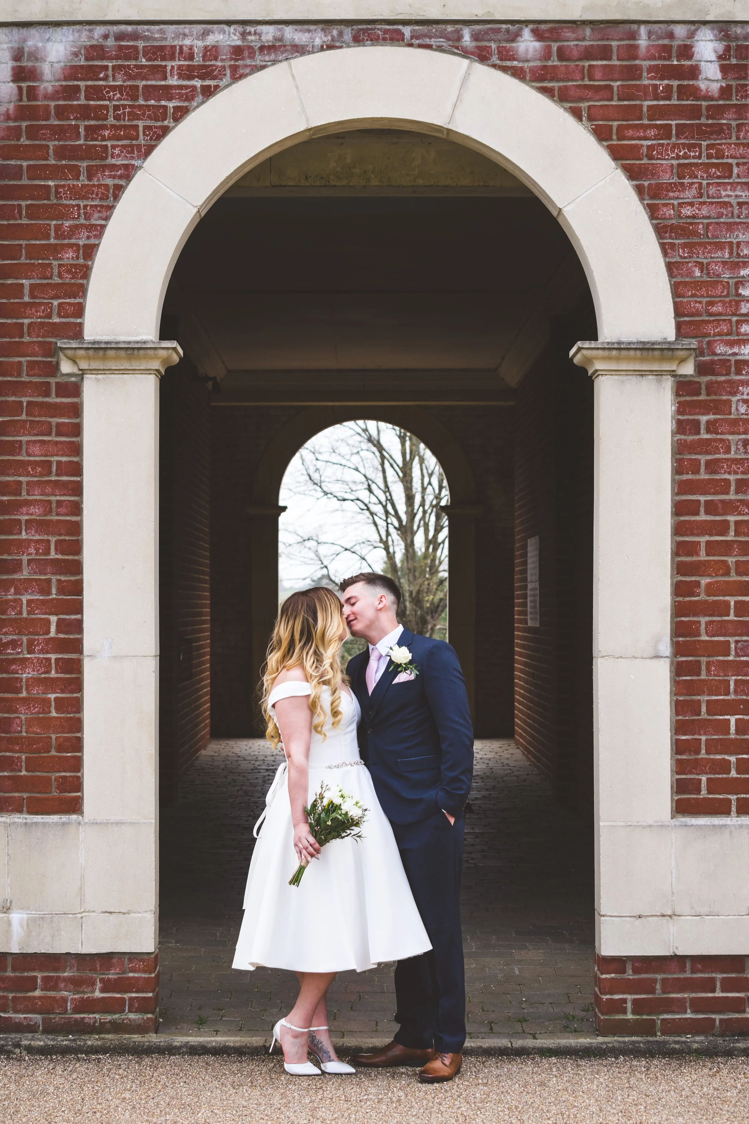 A bride and groom sharing a kiss under an arched brick walkway, with the bride holding a flower bouquet and both dressed in wedding attire.