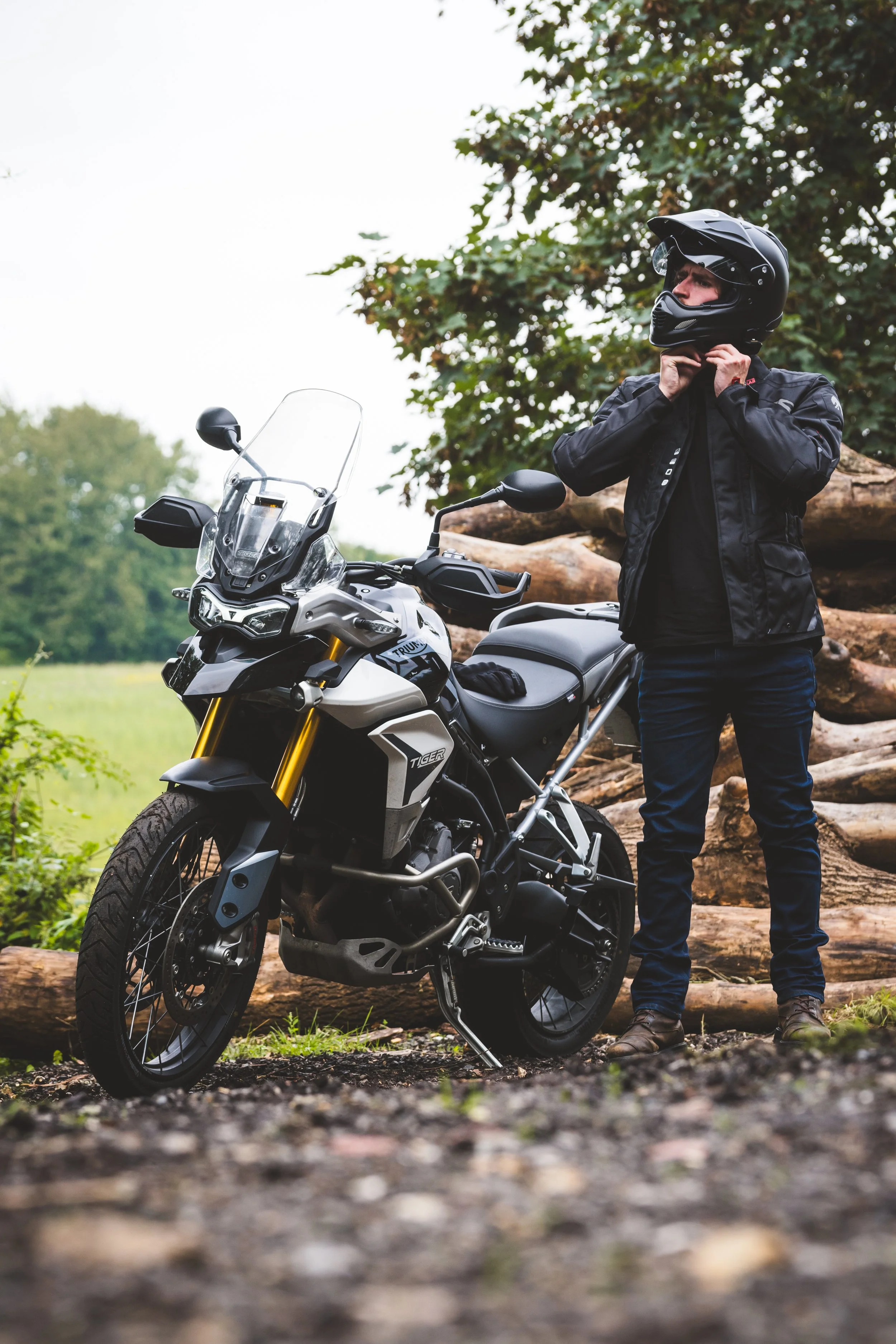A person wearing a black helmet and jacket stands outdoors next to a gray adventure motorcycle, adjusting their helmet strap, with trees and logs in the background.