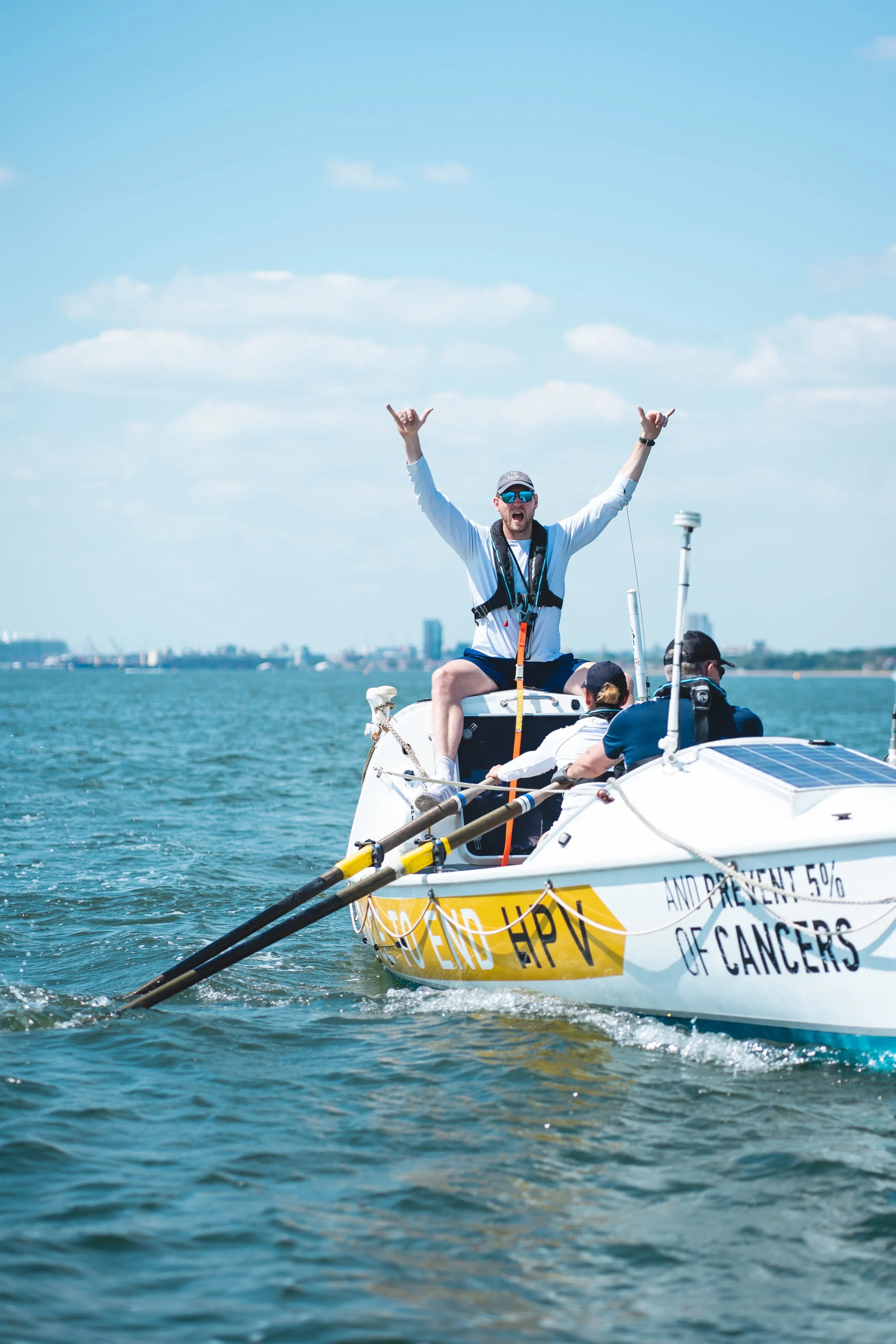 A group of people on a sailboat with a man standing at the front raising his arms in celebration on a sunny day with a city skyline in the background.