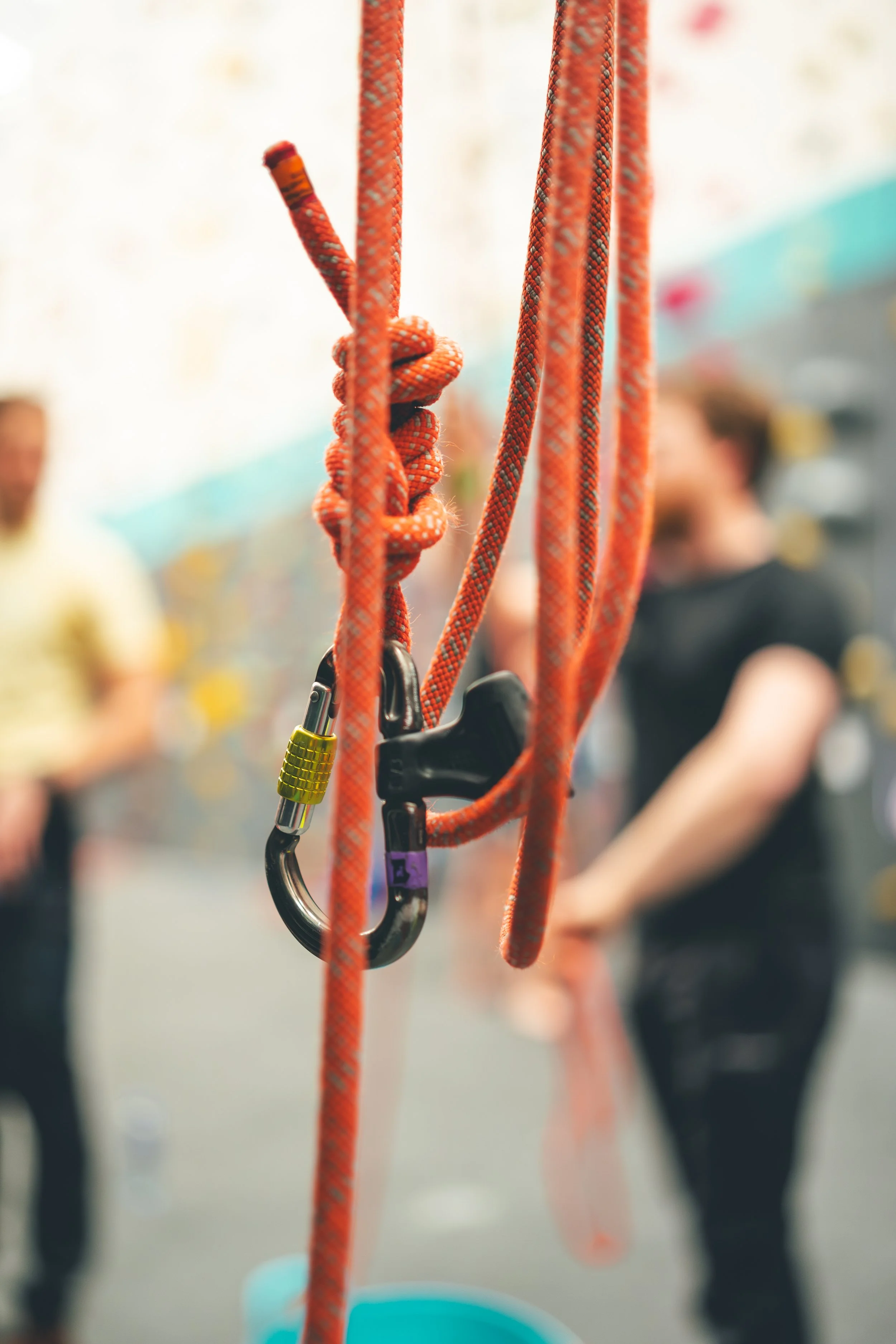 Climbing gym with orange climbing ropes and quickdraws, with blurred people in the background.