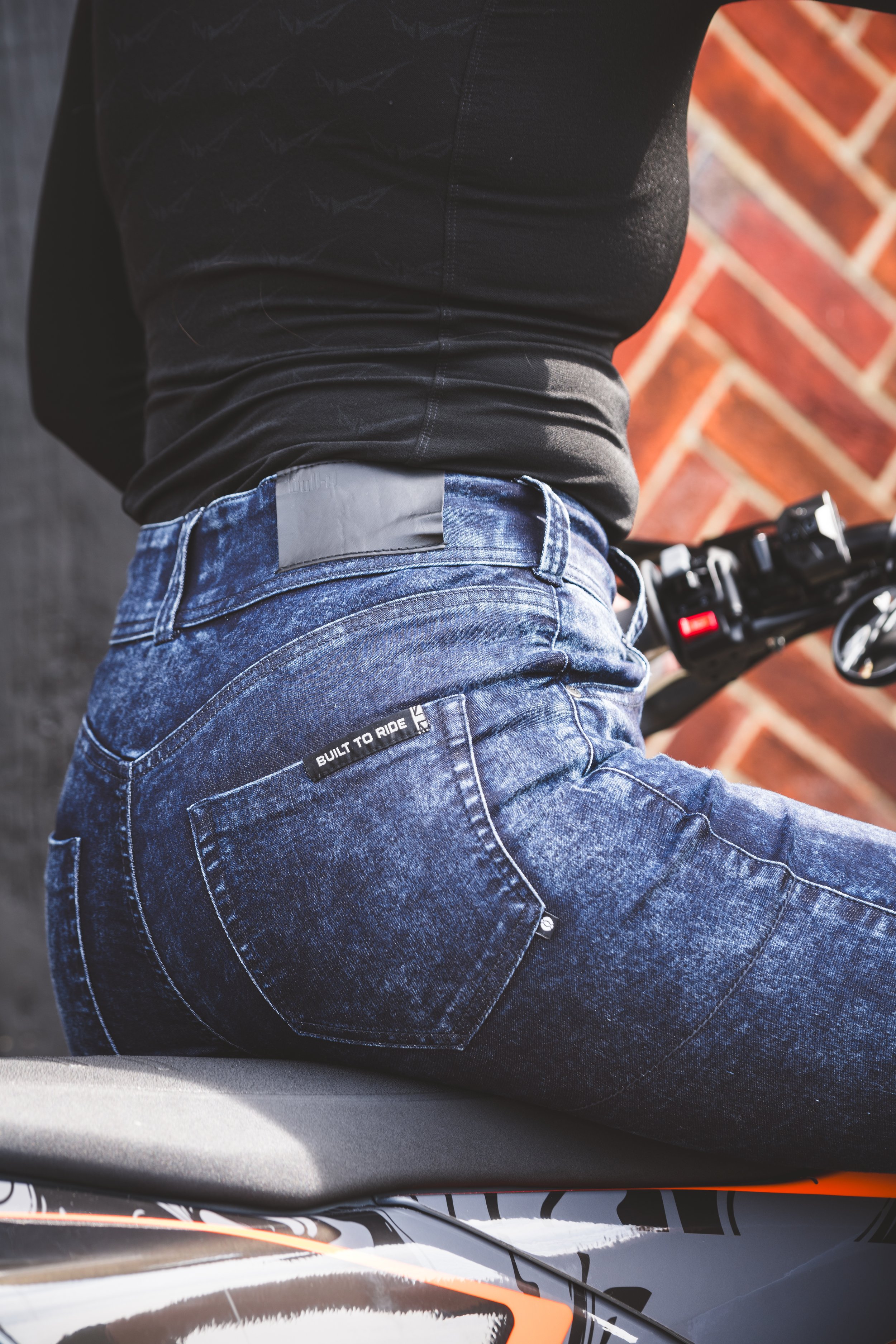 Close-up of a person wearing blue jeans and a black long-sleeve shirt seated on a motorcycle against a brick wall background.