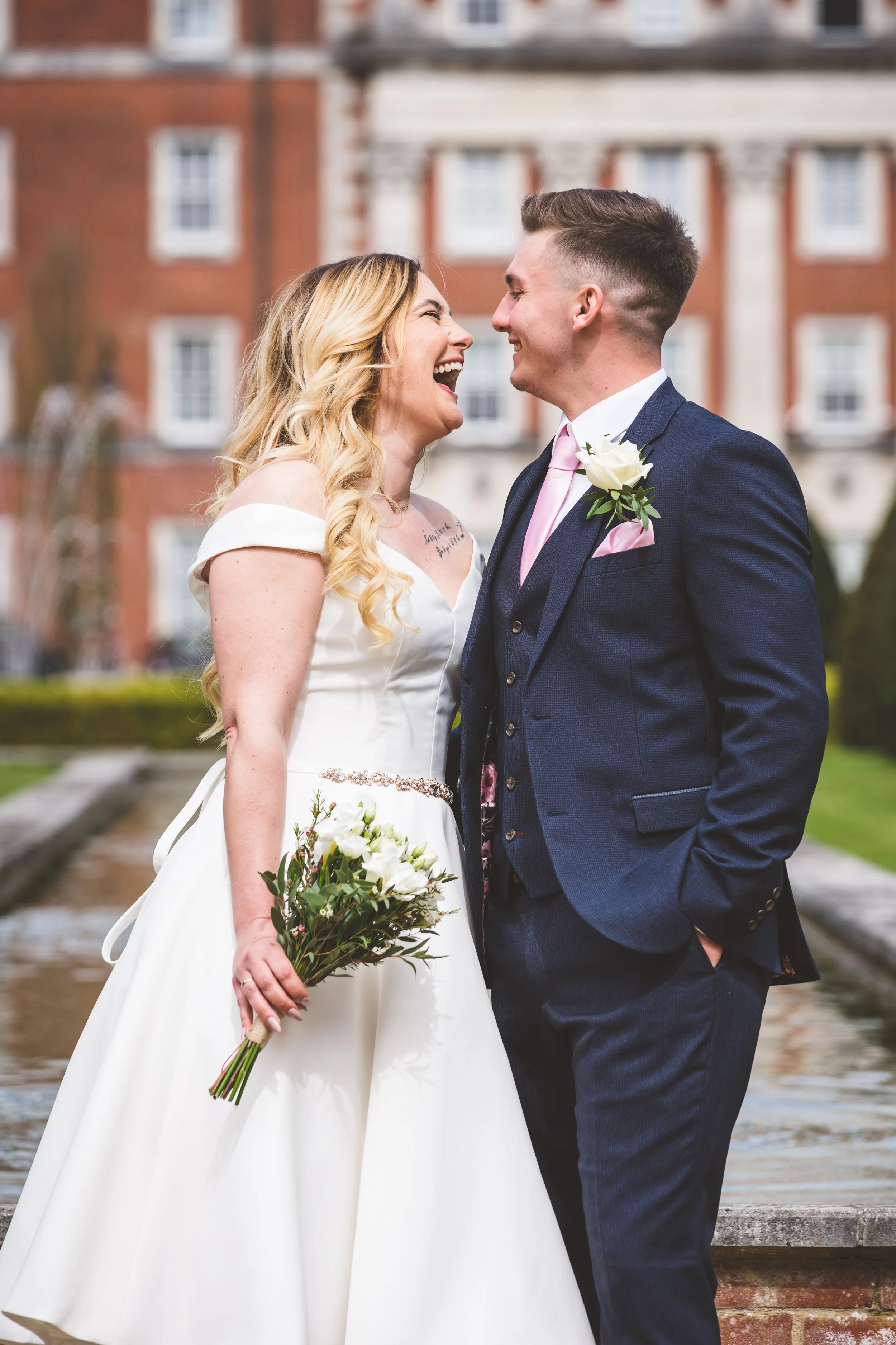 A smiling newlywed couple laughing and leaning toward each other outdoors in front of a historic building. The bride is holding a bouquet of white flowers, wearing a white off-shoulder wedding dress, and the groom is dressed in a navy suit with a pin