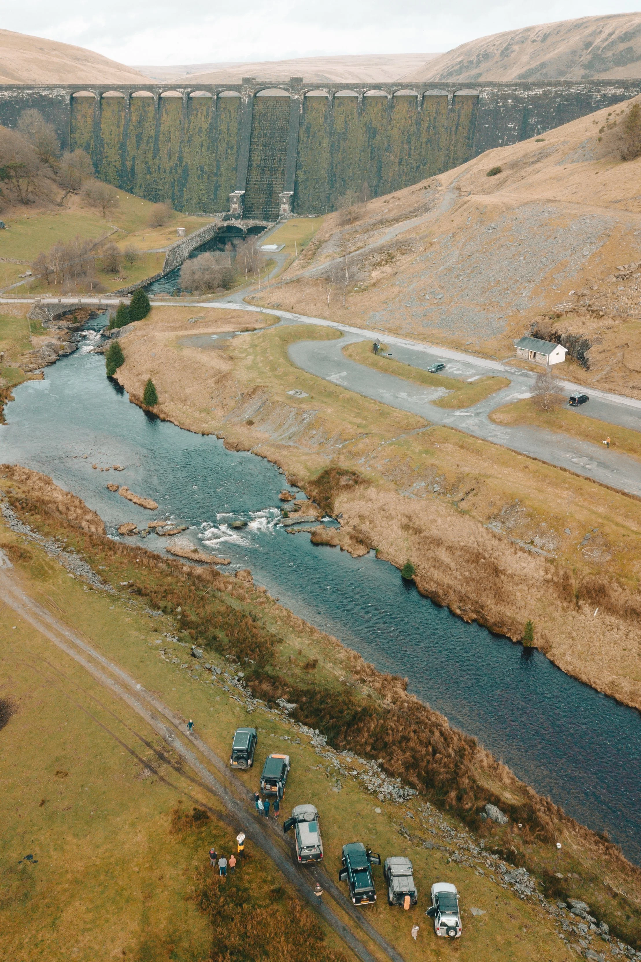 Aerial view of a dam in a rural, hilly landscape with a river flowing in the foreground. Several parked cars and a small building are near the riverbank, with a group of people nearby. The dam spans across the hills in the background.