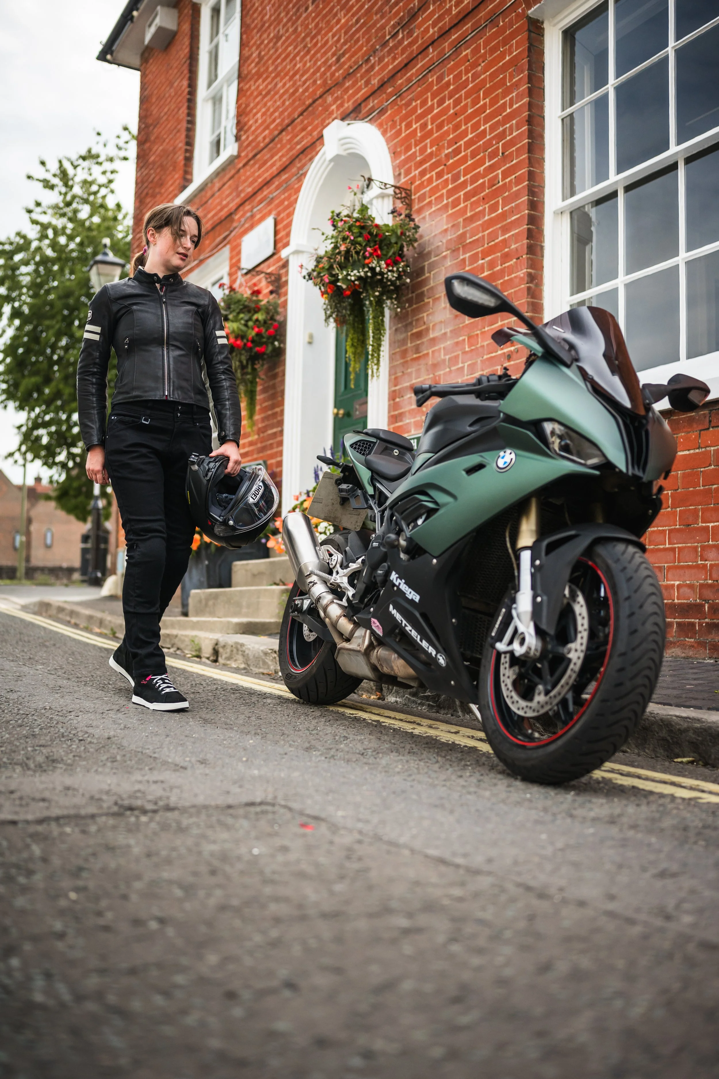 A young woman dressed in black motorcycle gear stands on a city street holding a helmet, looking at a green BMW motorcycle parked on the side of the road in front of a brick building with flower baskets.