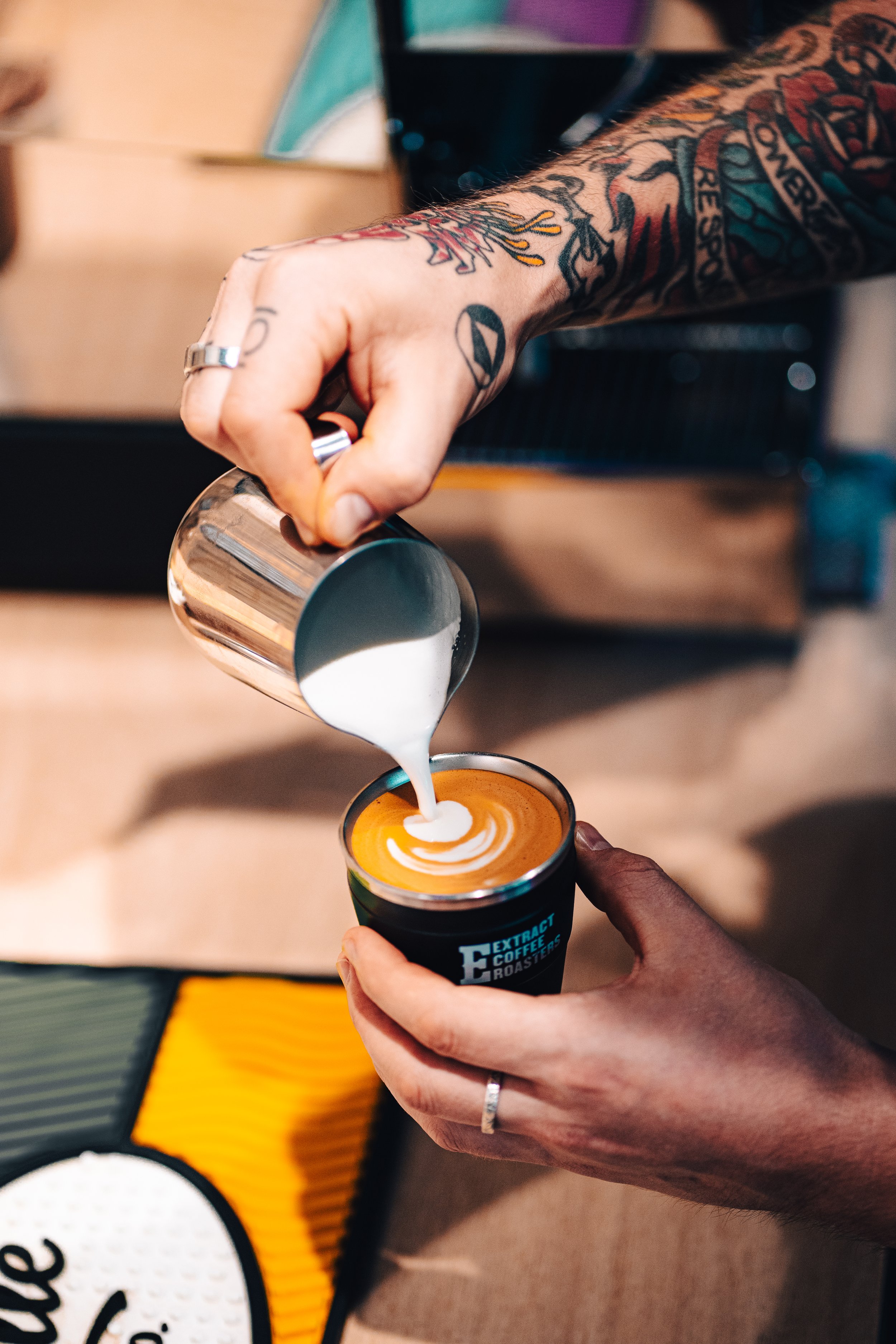A person with tattooed arms is pouring steamed milk from a small pitcher into a cup of coffee, creating latte art. The coffee cup has the logo 'Extract Coffee Roasters'.