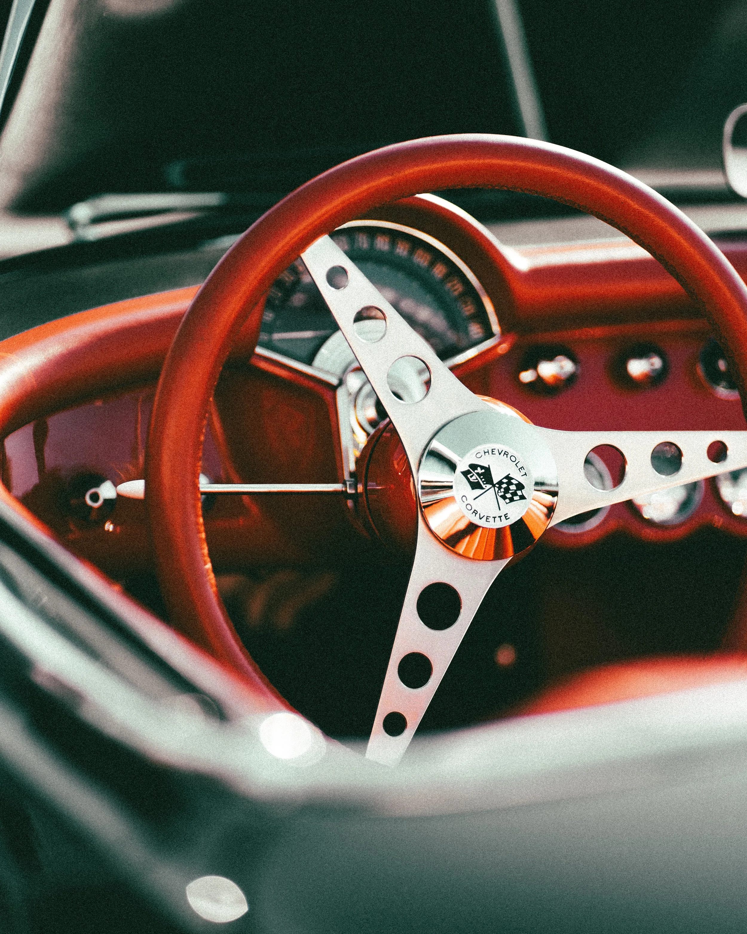 Close-up of a vintage car's interior, highlighting a Chevrolet Corvette steering wheel with a red rim and metal spokes, and a dashboard with a speedometer and gauges.