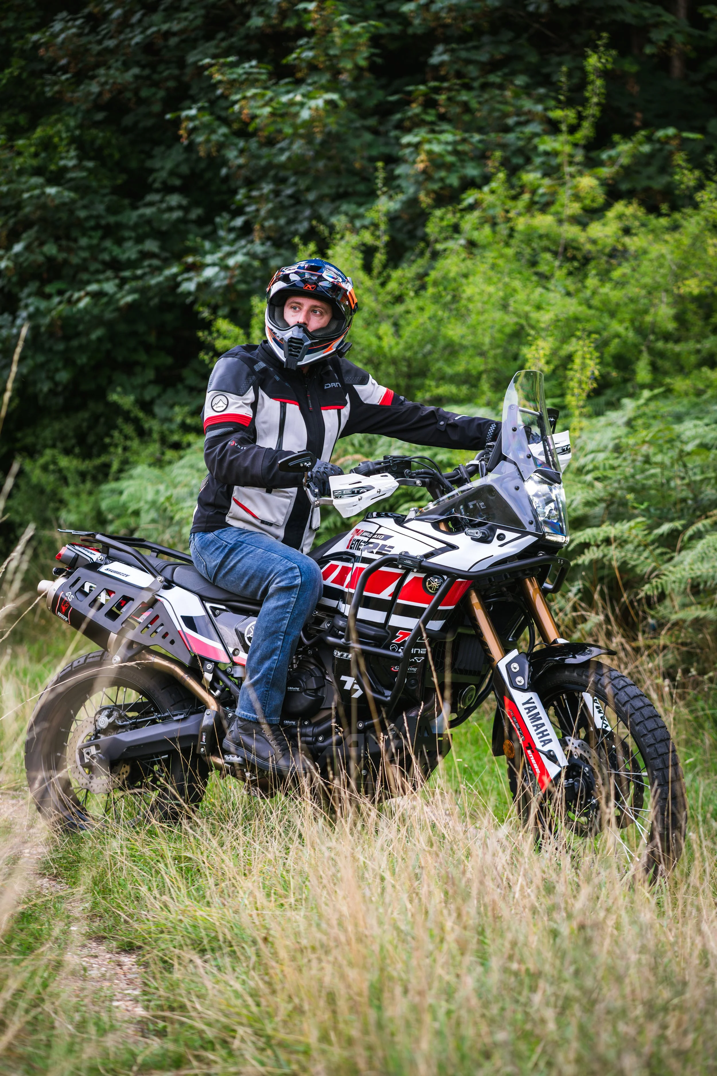 A person wearing motorcycle gear and a helmet, sitting on a Yamaha off-road motorcycle in a grassy outdoor setting with green trees in the background.